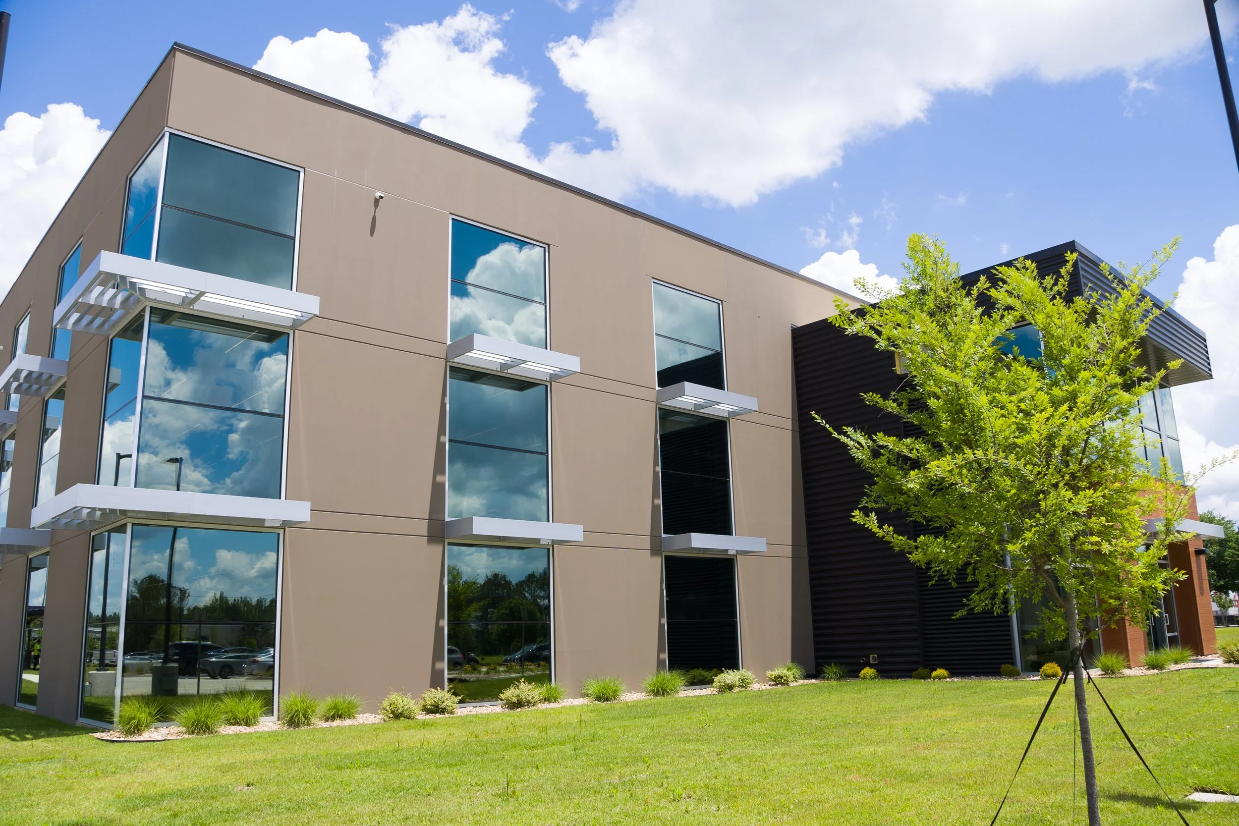 Modern multi-story office building with large reflective windows, a green lawn in the foreground, a young tree, and a bright blue sky with scattered white clouds.