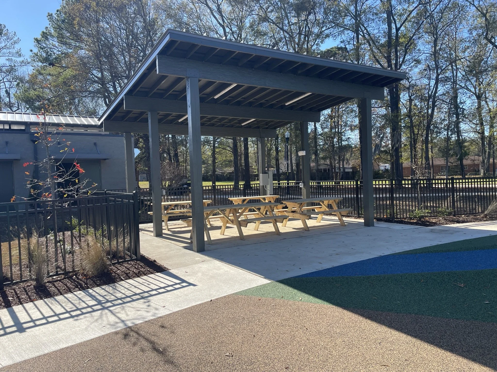 An outdoor pavilion with a roof and picnic tables underneath, surrounded by a black metal fence and trees with autumn foliage.