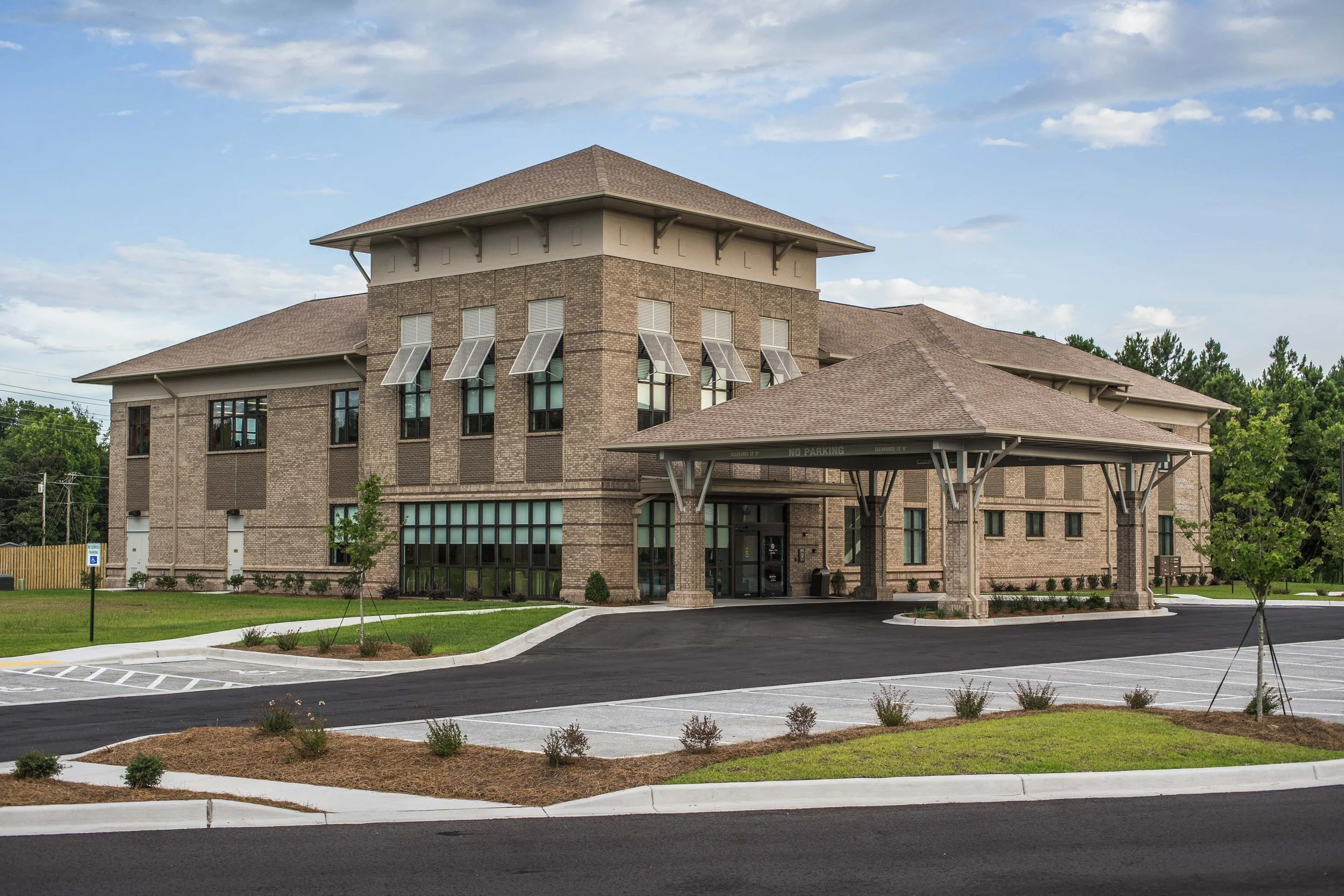 A brick building with large windows and an overhanging roof, surrounded by a parking lot, small trees, and landscaped areas, on a partly cloudy day.