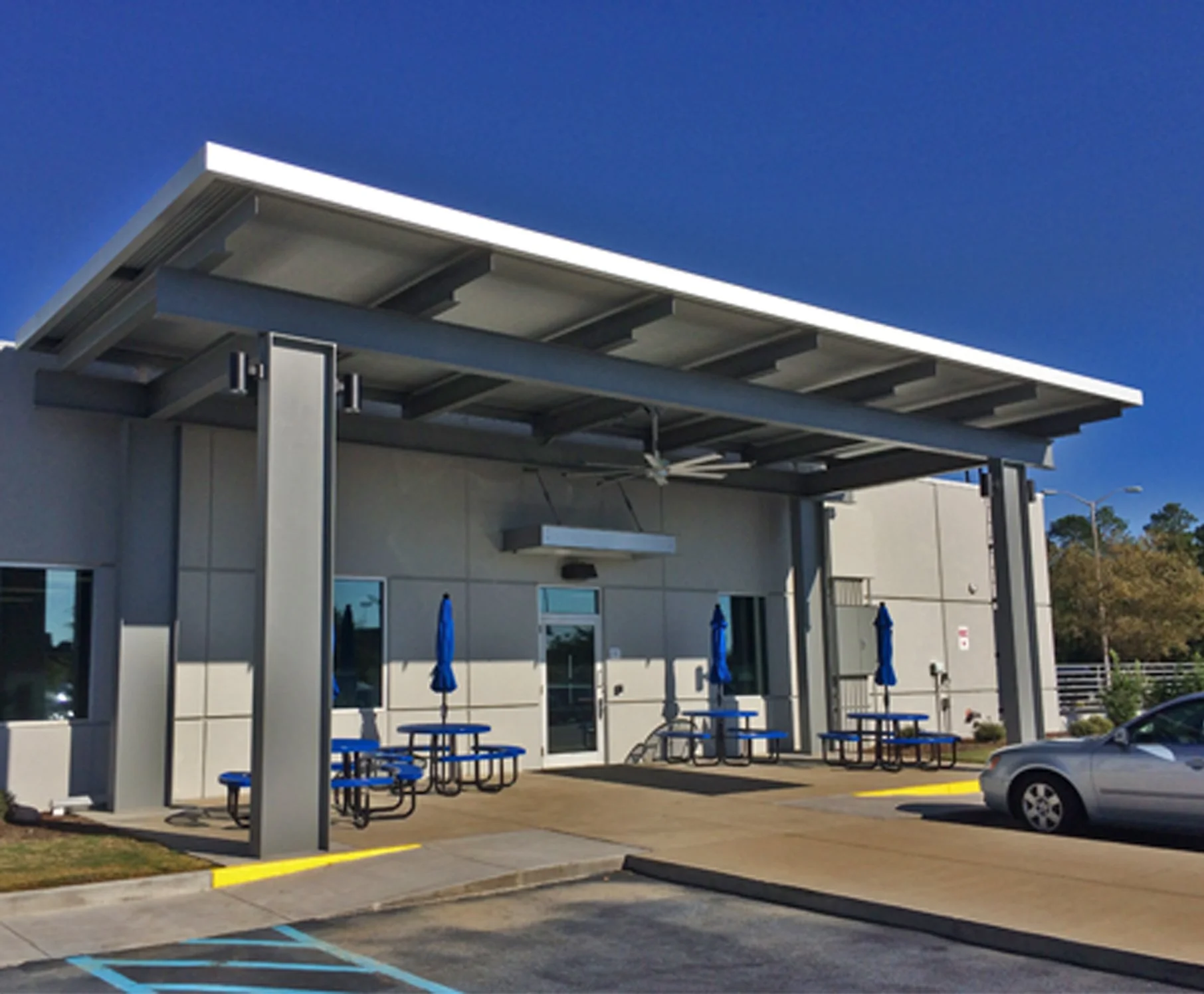 Exterior view of a restaurant patio covered by a modern metal roof with blue tables and chairs, blue umbrellas, and a parking lot with marked spaces in front, under a clear blue sky.