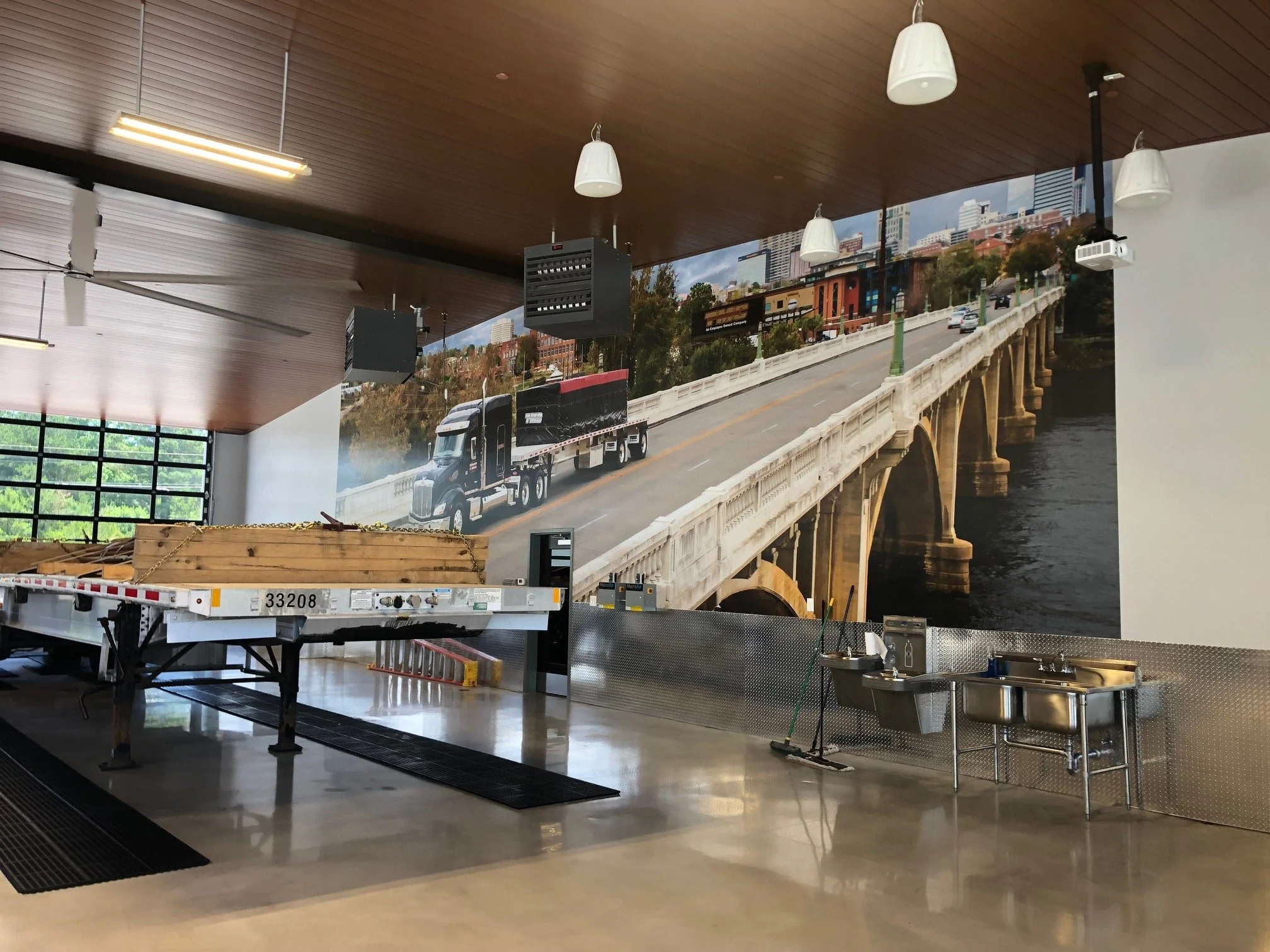Indoor space with a large wall mural of a cityscape and a bridge, featuring a truck on the bridge, with ceiling lights, a ceiling fan, some construction equipment, and a stainless steel sink area.