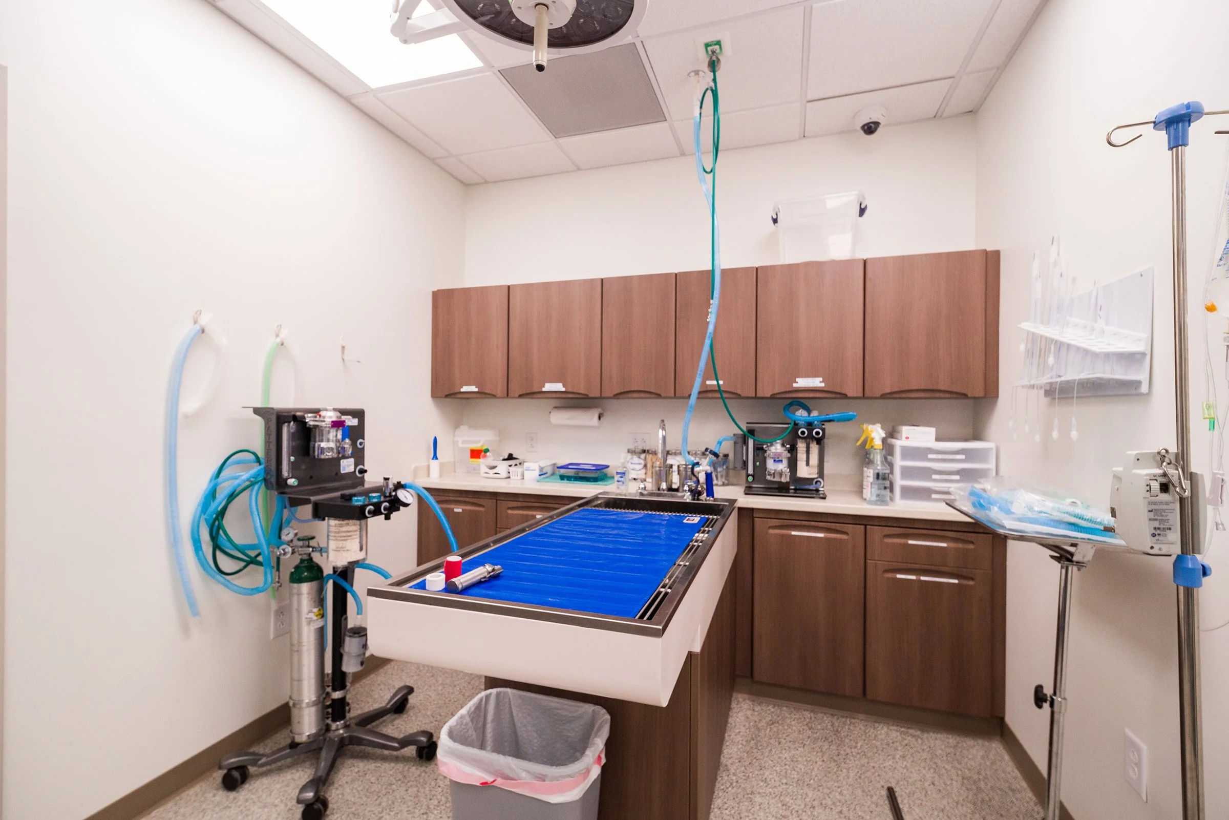 An operating room with medical equipment, including monitors, a surgical table, and supplies. The room has overhead lighting, wooden cabinets on the wall, and medical tubing hanging from the ceiling.