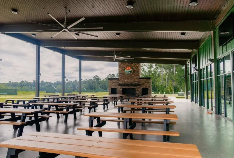 Empty outdoor pavilion with picnic tables, a large brick fireplace, and ceiling fans, overlooking a golf course and trees in the background.