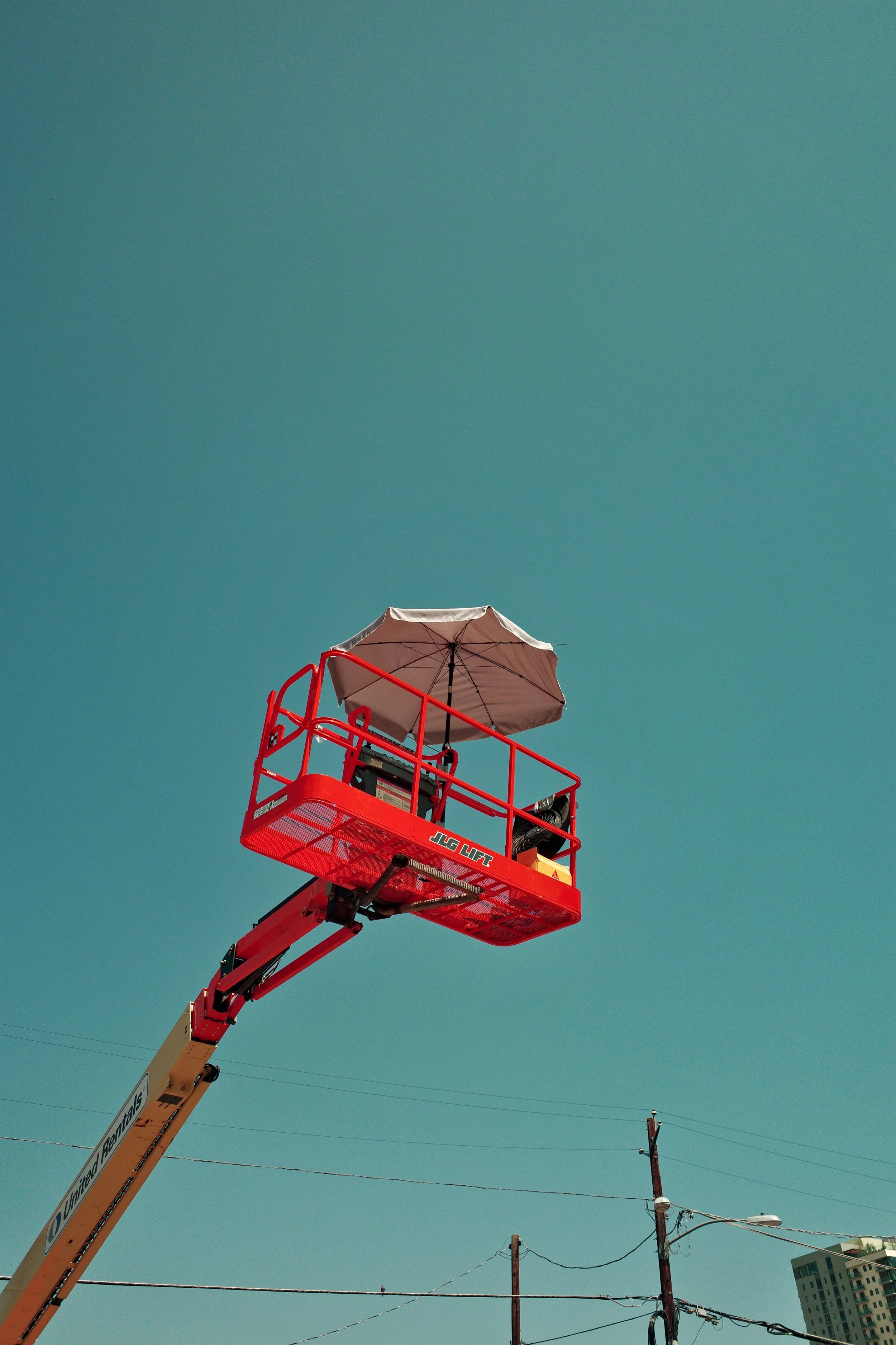 A raised red scissor lift with an umbrella attached to it, against a clear blue sky, with power lines and a building in the background.