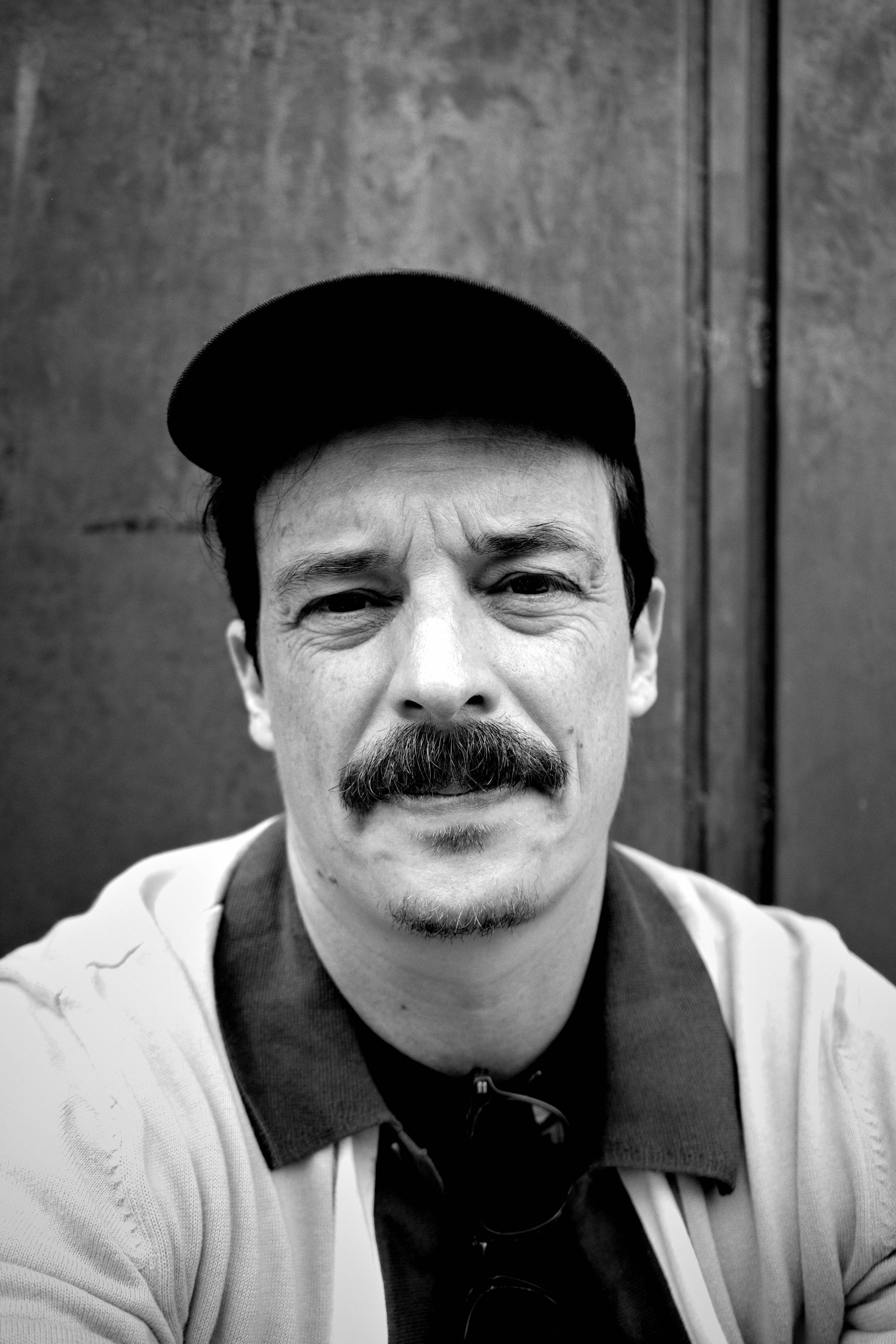 Black and white photo of a man wearing a baseball cap, mustache, and goatee, smiling slightly at the camera, with a plain background.