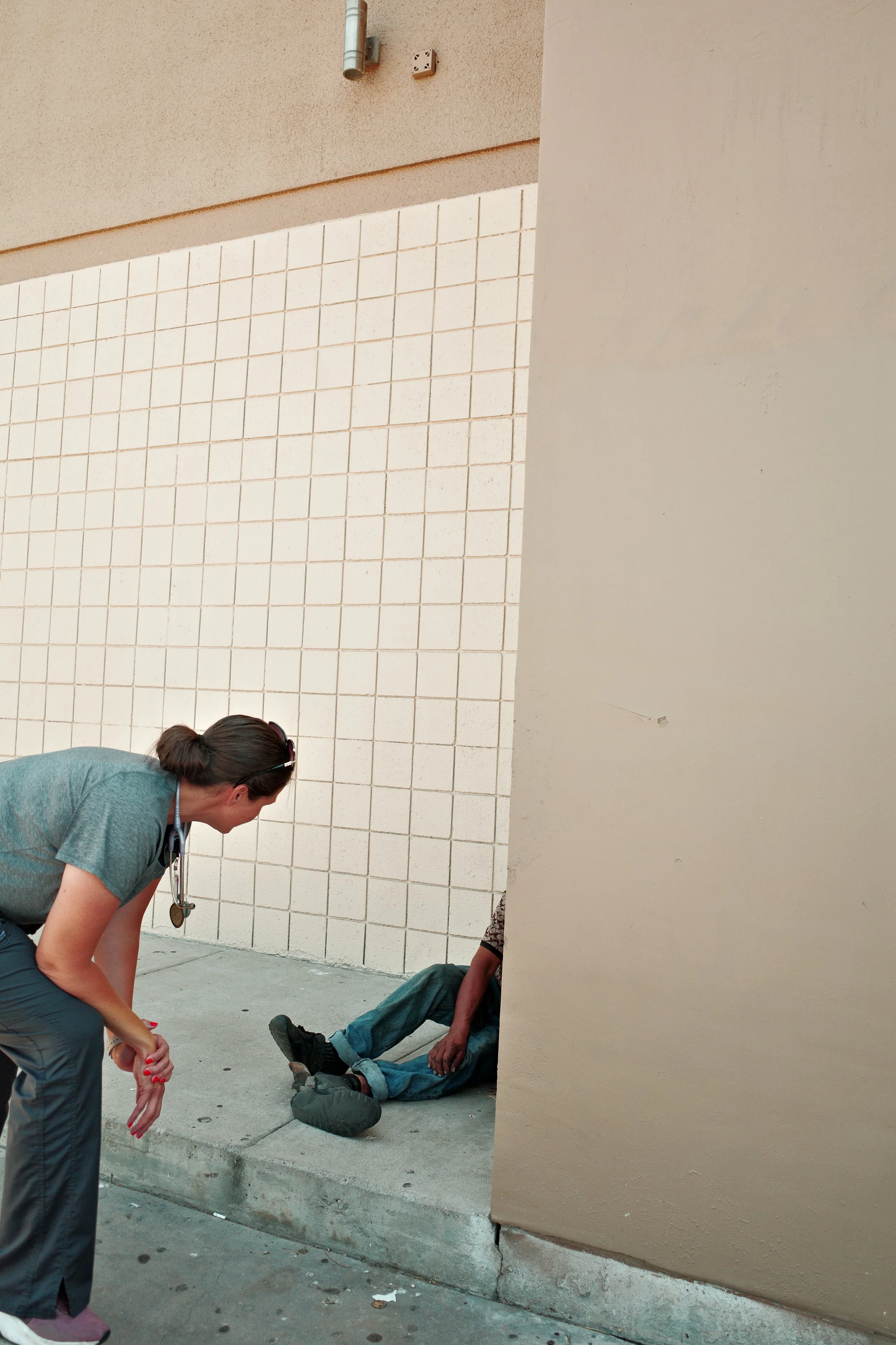 A person lying on the ground with their eyes closed, and another person kneeling beside them, appears to be performing first aid. It is outside, against a beige wall with a tiled section and a sidewalk.