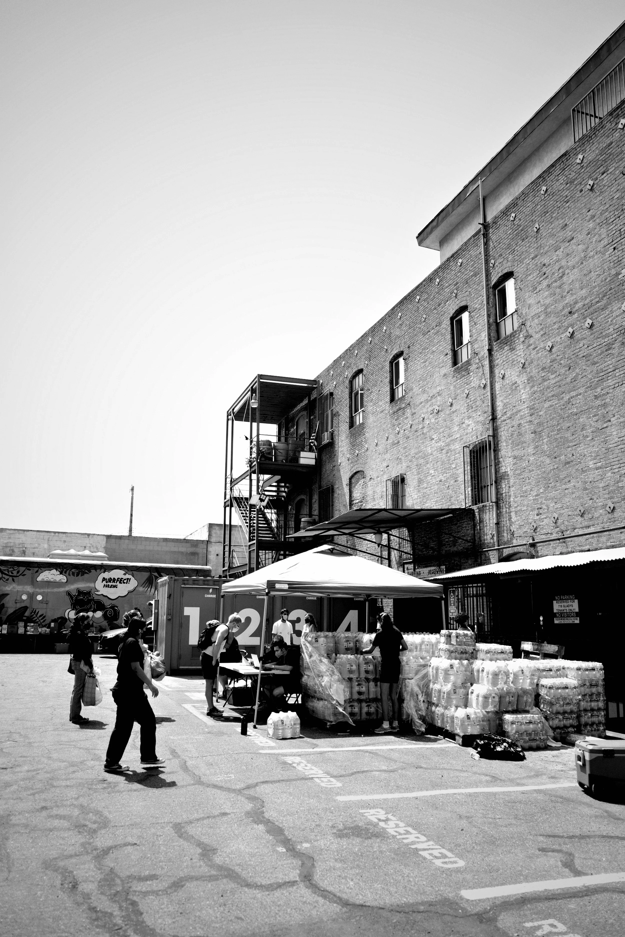 People shopping for bottled water at an outdoor market, with a brick building in the background and parking spaces marked on the pavement.