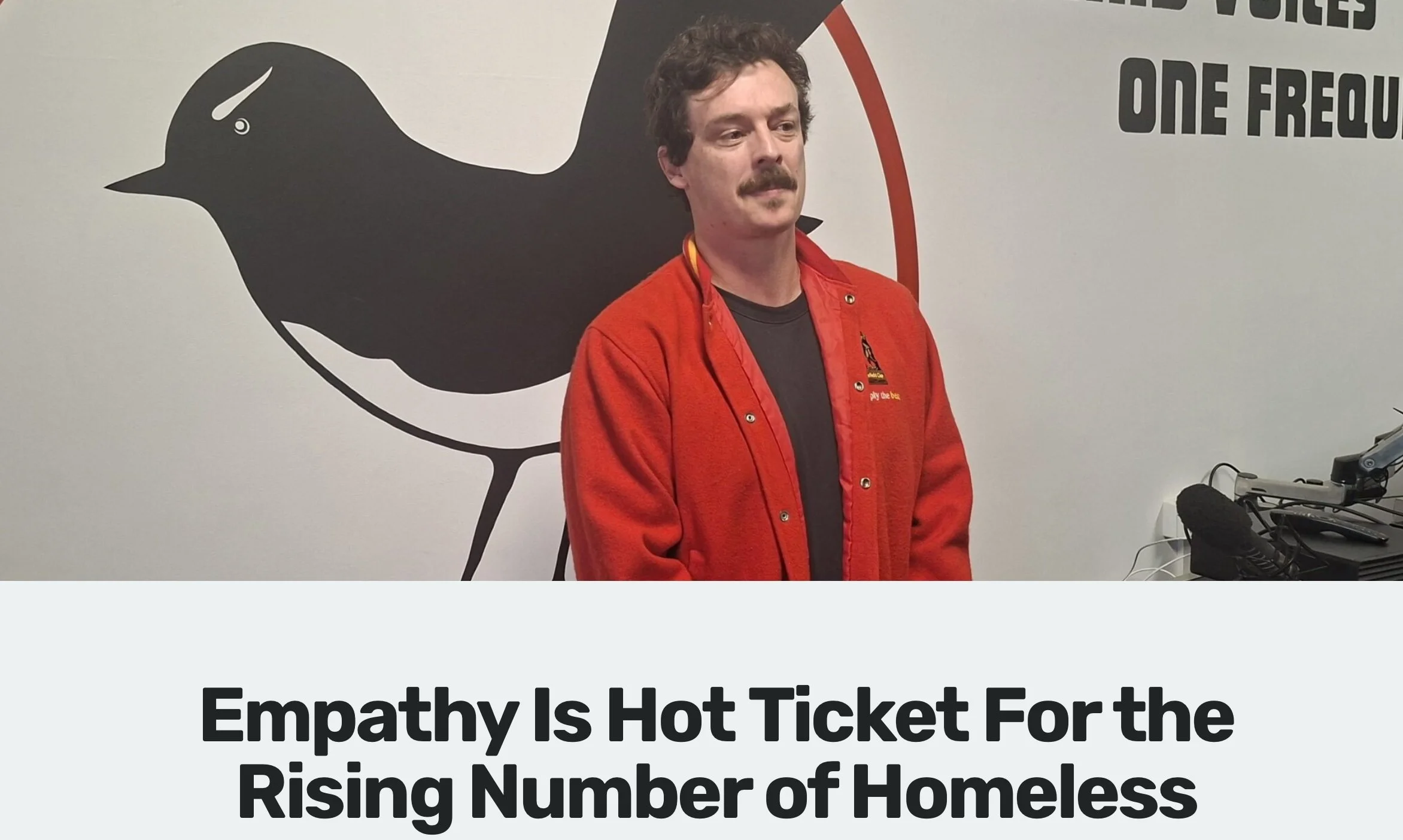 A man with a mustache and curly hair wearing a red jacket standing in front of a wall with a large bird graphic and part of a sign that says 'One Flew...' and a smaller text below that is not fully visible.