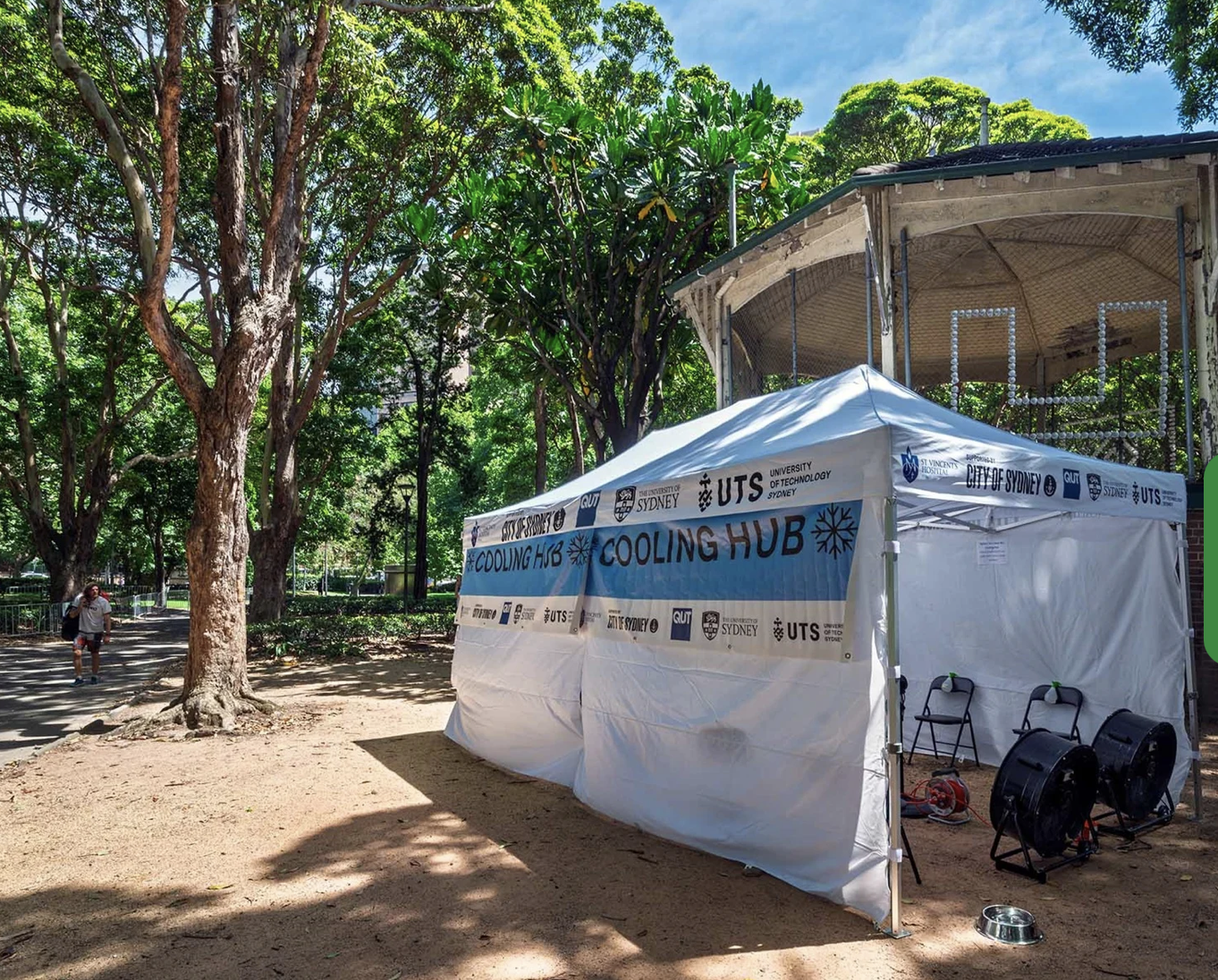 A white tent with a blue banner reading 'Cooling Hub' set up outdoors near large trees, with two black fans in front and a small person walking on the path on the left side of the image.