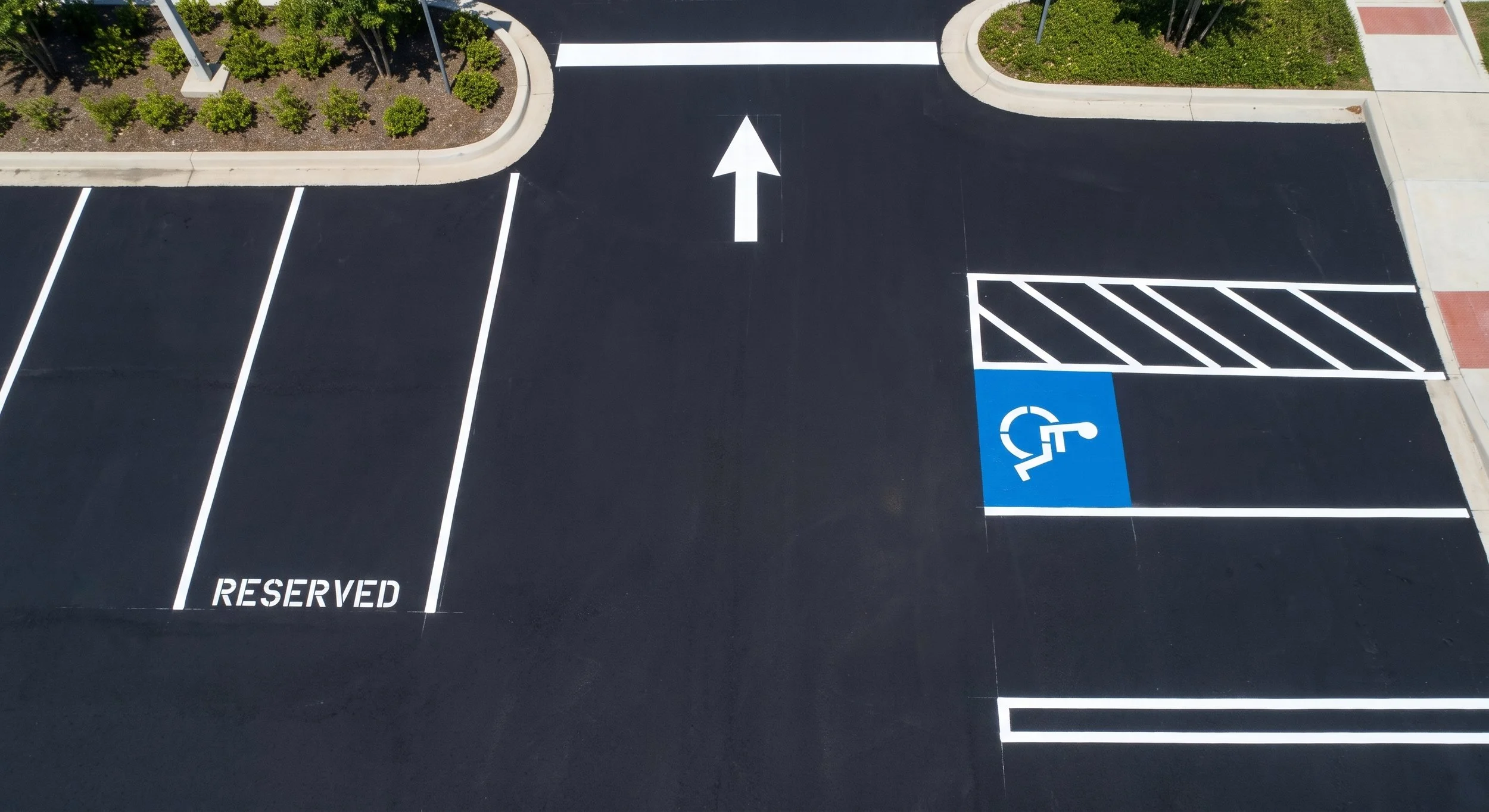 Empty parking lot with a reserved space for disabled parking marked by a blue sign and painted lines, a white arrow directing forward, and a striped section indicating a no-parking zone.