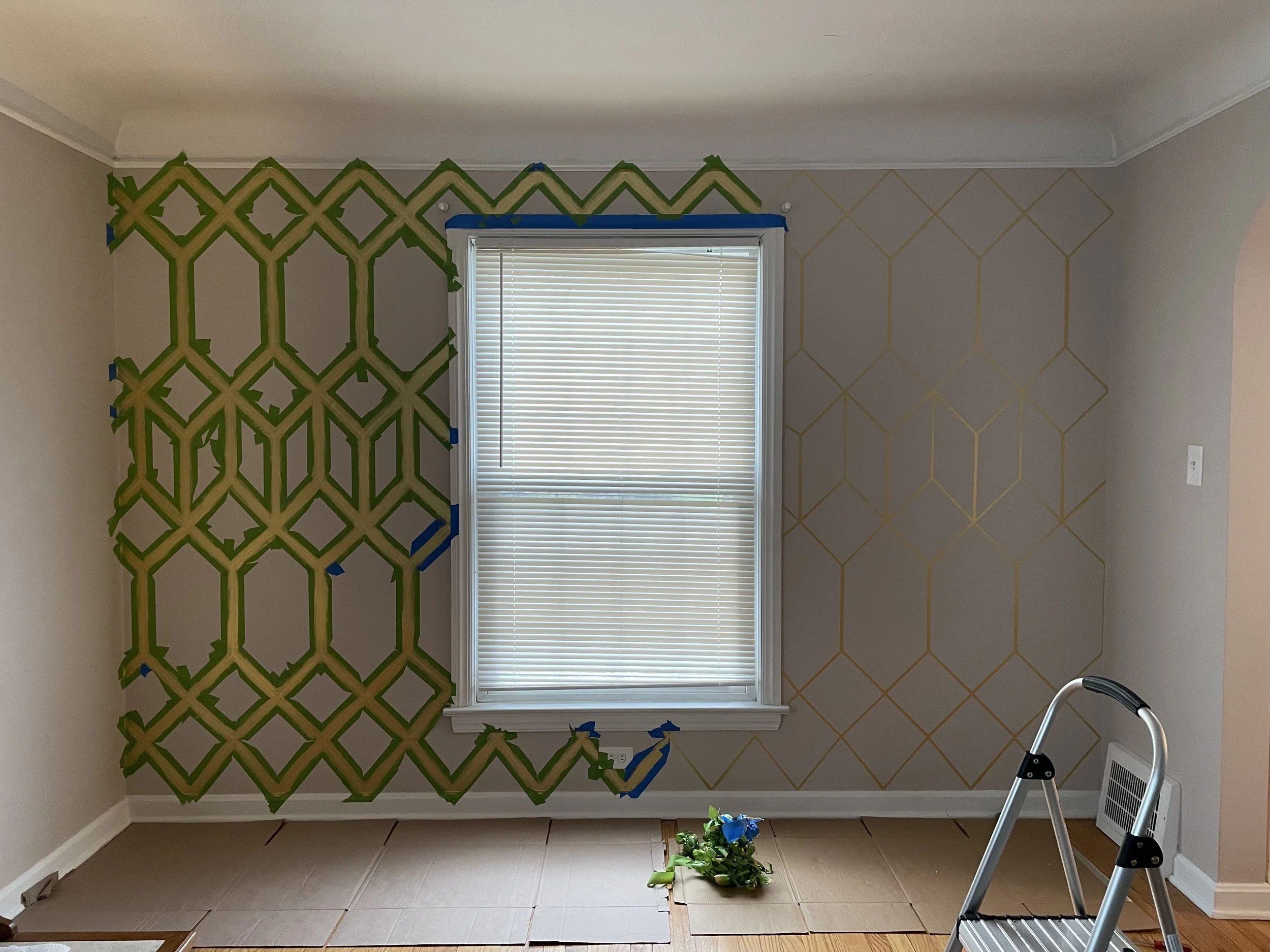 Room with a window covered by blinds, walls partially painted with painter's tape and green paint lines, and a small plant on the floor. A step ladder is visible on the right side.