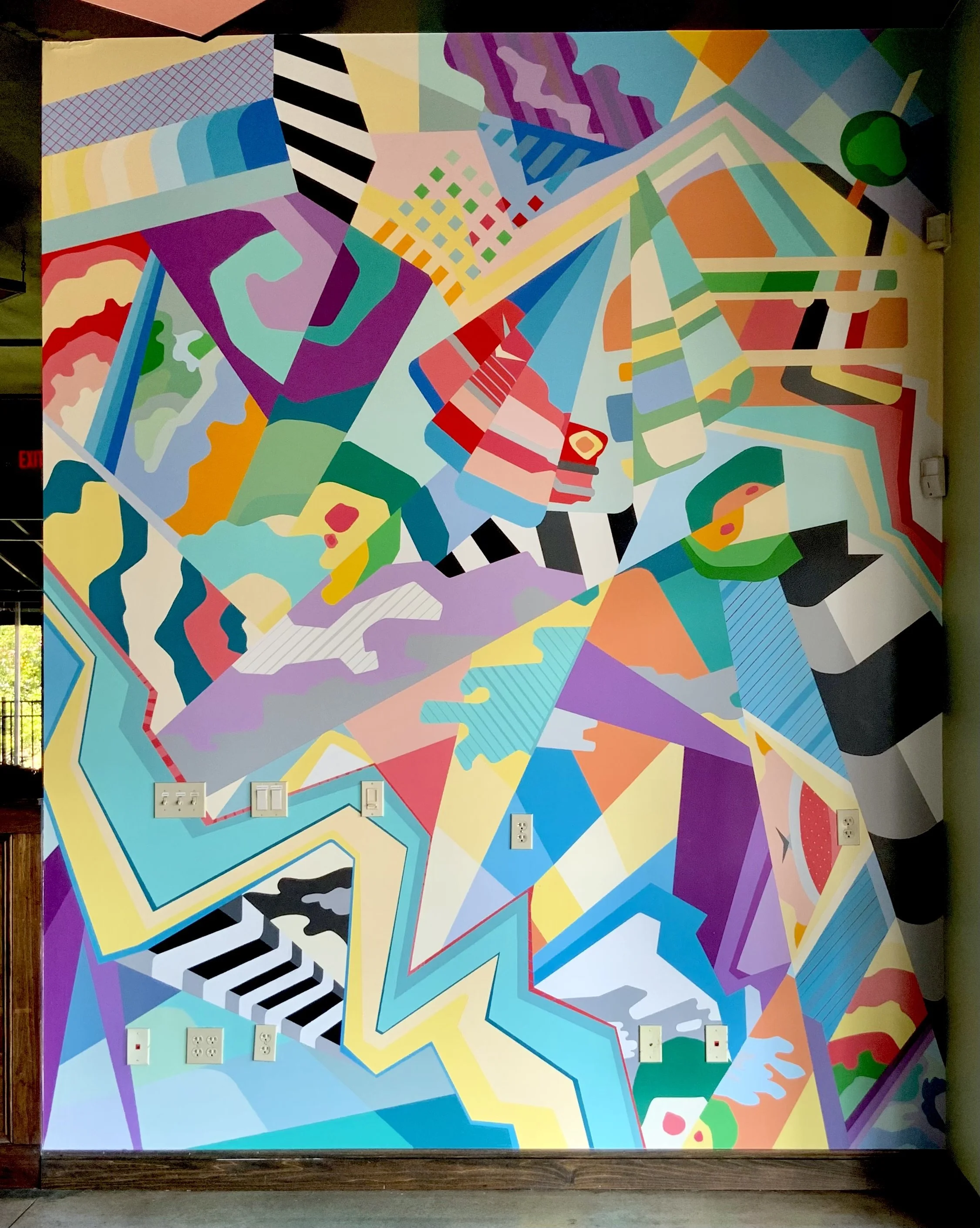 Colorful abstract mural with geometric shapes and patterns, featuring various bright and pastel colors, covering a wall with electrical outlets and switches.