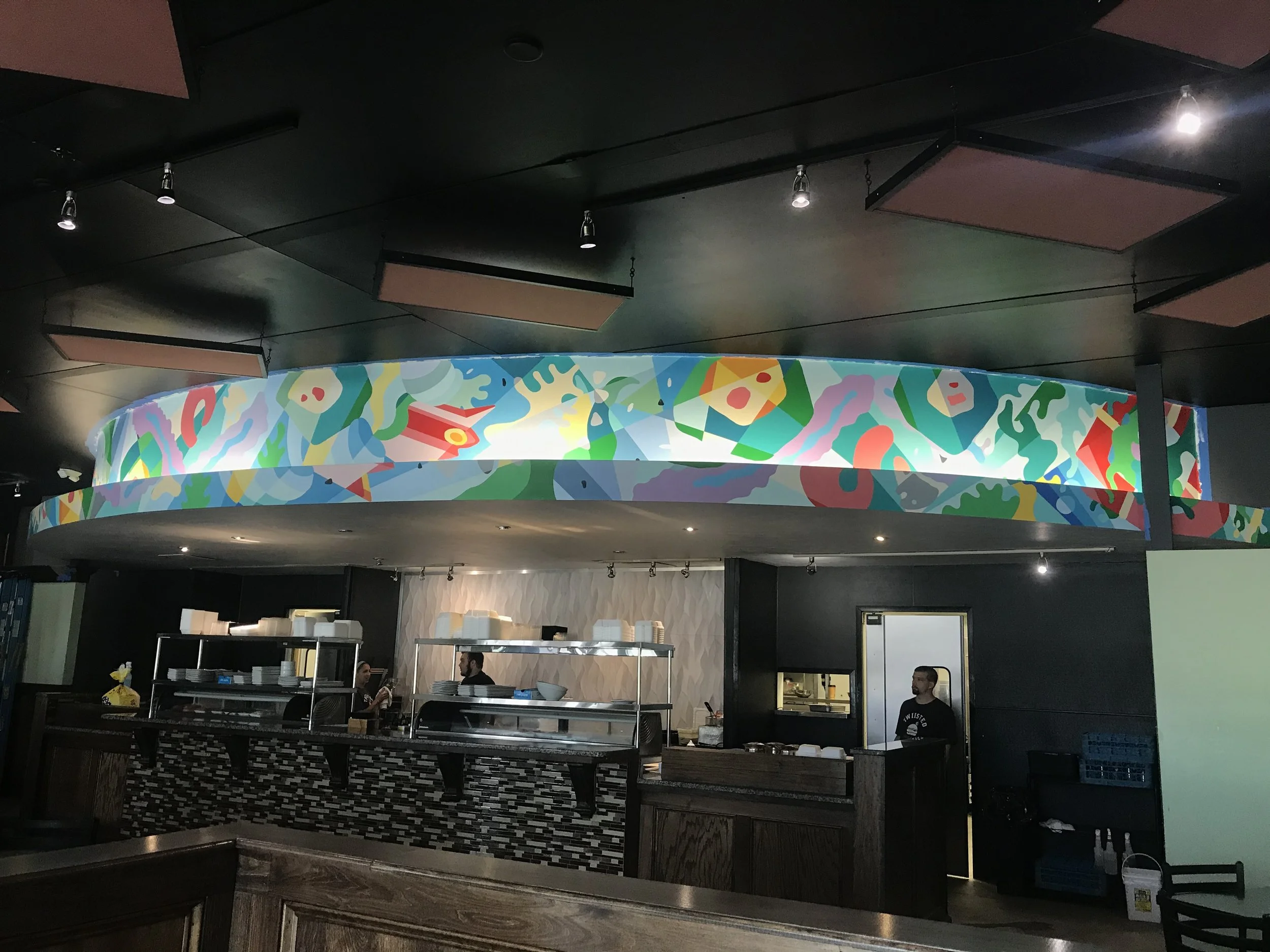 Interior of a restaurant or café with a dark ceiling featuring hanging black panels and spotlights. A colorful, abstract circular artwork or decoration hangs above a counter or serving area where people are standing and talking.