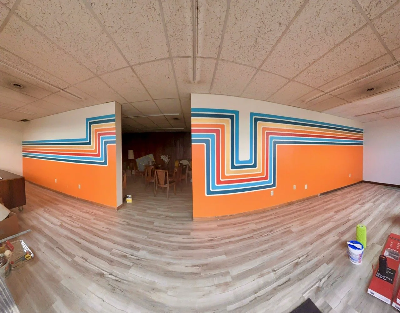 Interior of a room with a colorful, retro-style stripe mural on divided walls featuring orange, blue, yellow, and white colors, light wood flooring, and a partially visible area with chairs and a dark wooden wall.
