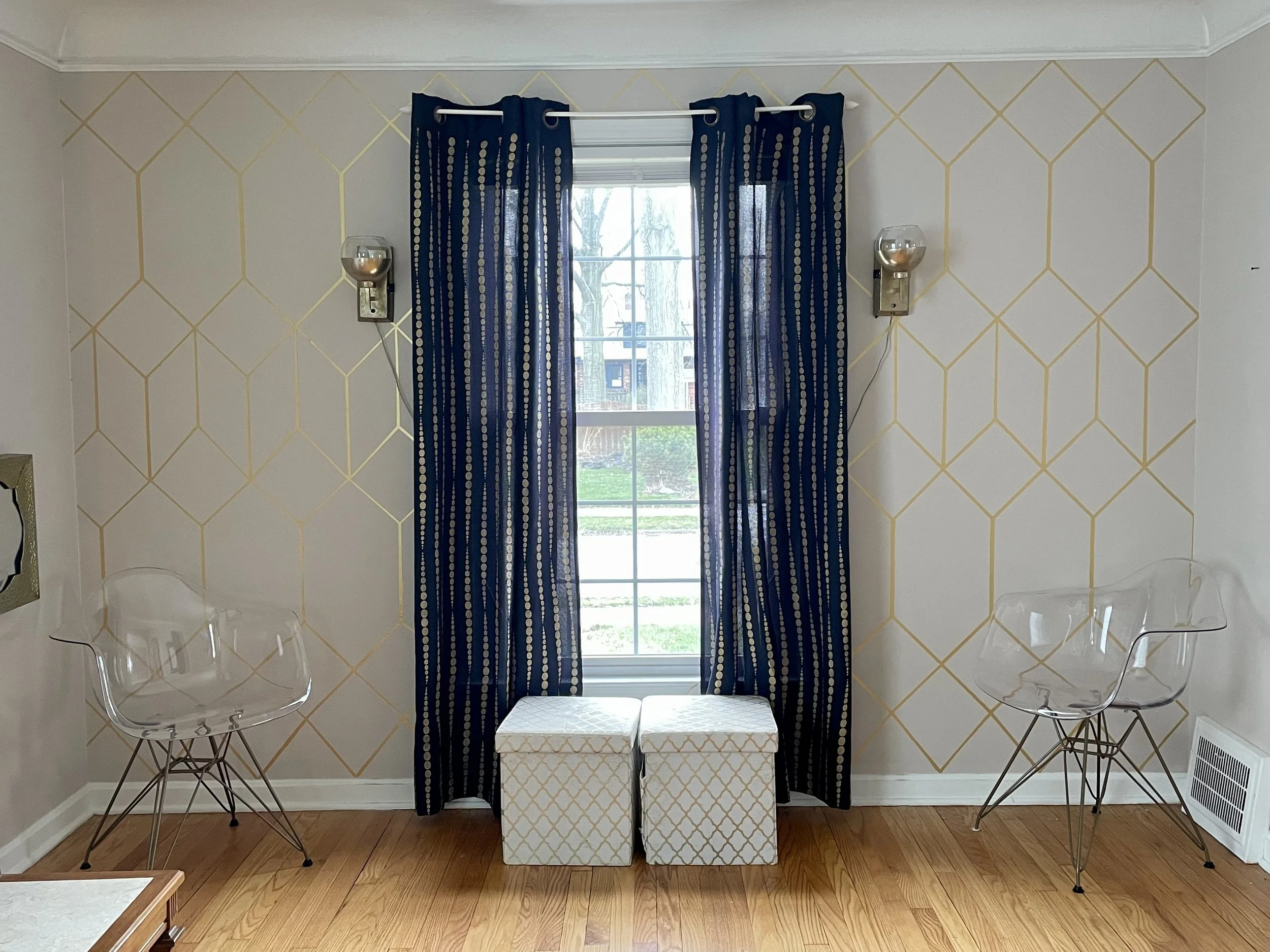 A living room with decorated wall, window with blue striped curtains, two transparent chairs, and a patterned box in front of the window.