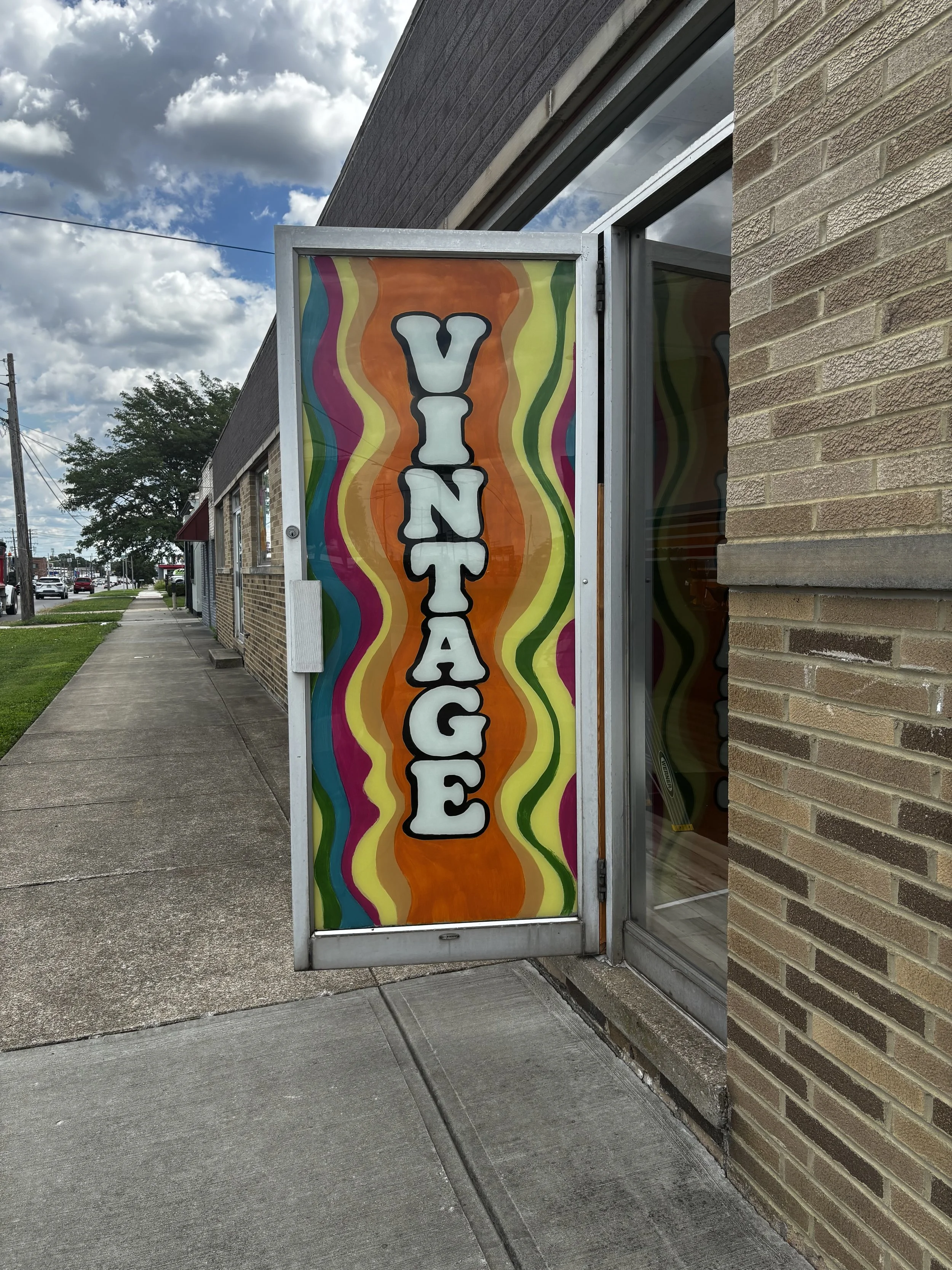 Colorful sign with the word "VINTAGE" written vertically in bold black and white letters, with vibrant wavy rainbow stripes background, attached to the side of a brick building on a sidewalk.
