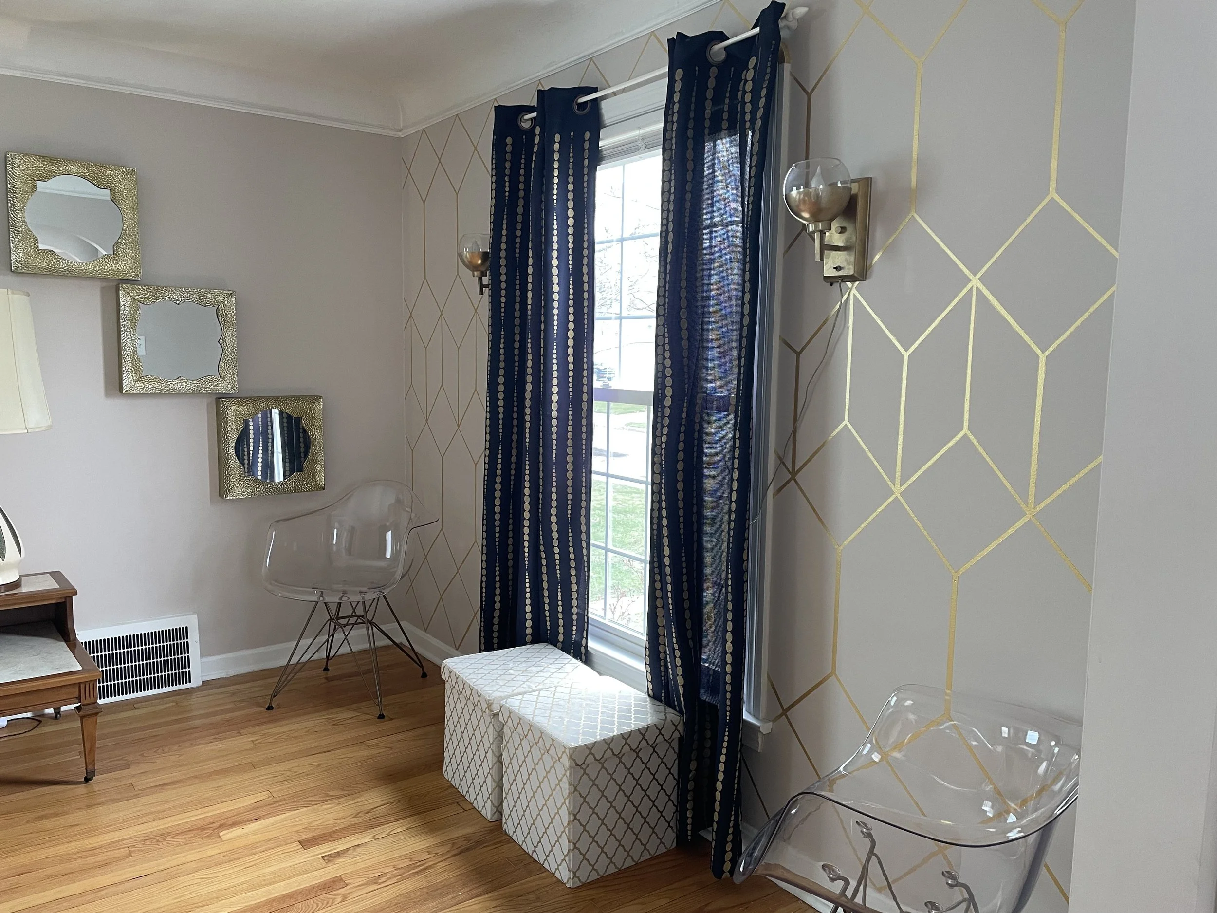A corner of a living room with a window dressed in dark blue curtains with white circular patterns, two wall-mounted candle-style lights, and decor mirrors on the wall. There is a clear acrylic chair, a dark wooden side table, a white storage bench w
