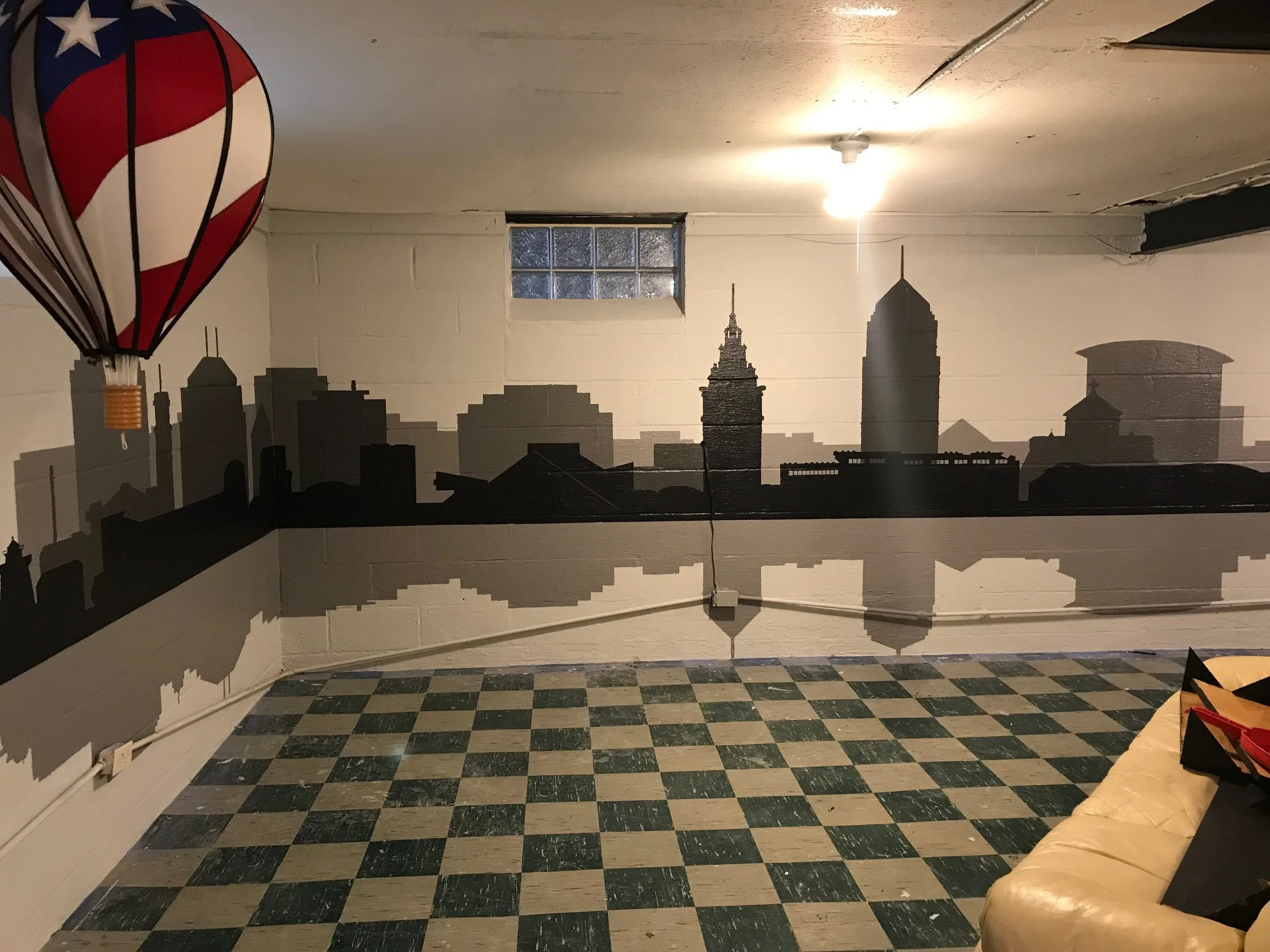 A room with a wall decorated with a city skyline silhouette mural and a window. The ceiling has visible damage with peeling paint. The floor has a checkered pattern with green and beige tiles. There is a hanging lamp and a partial view of a beige sof