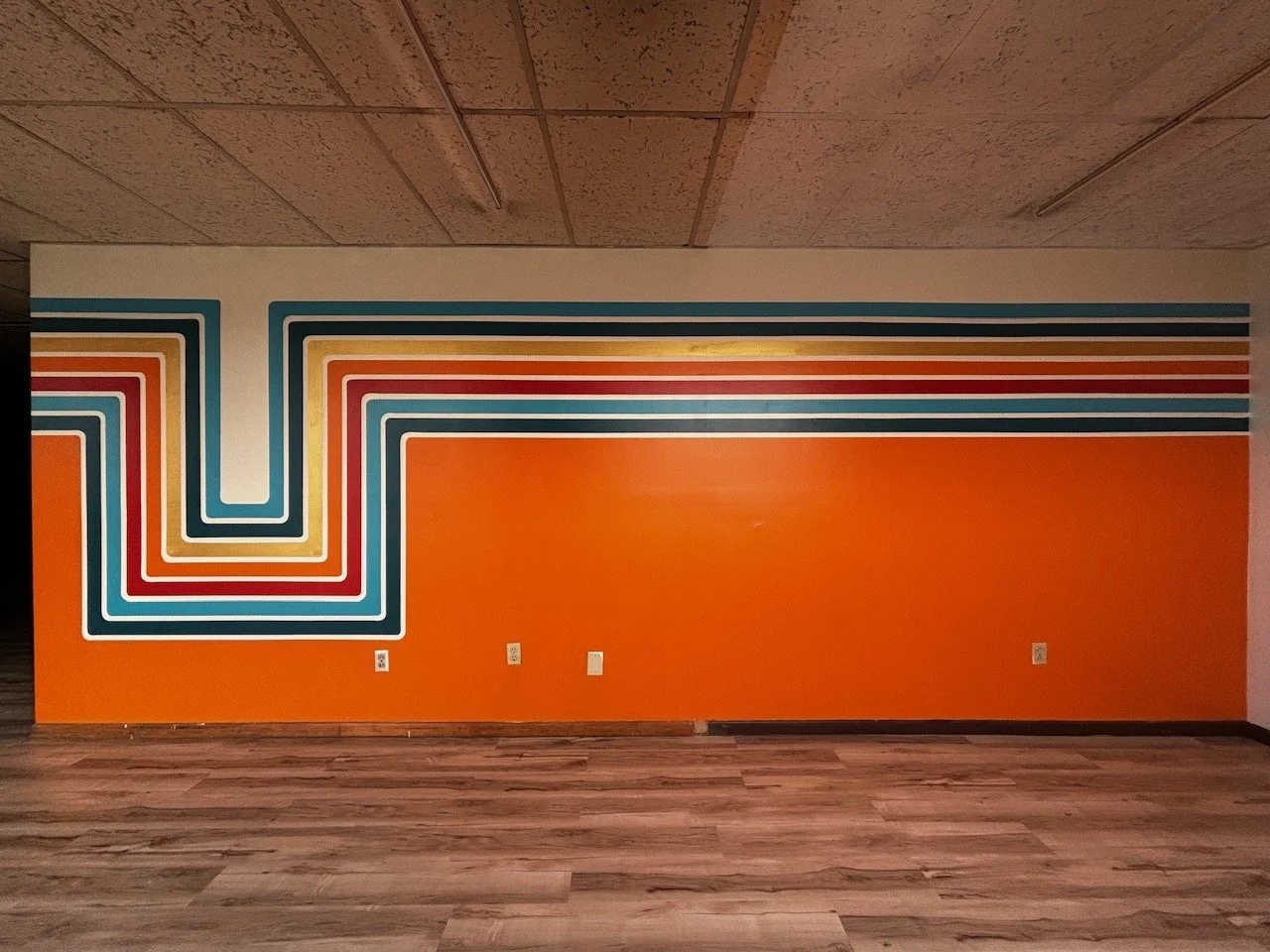 Colorful geometric mural with concentric lines in blue, red, yellow, white, and orange on an orange wall, with a wood-patterned floor and ceiling with linear lighting.
