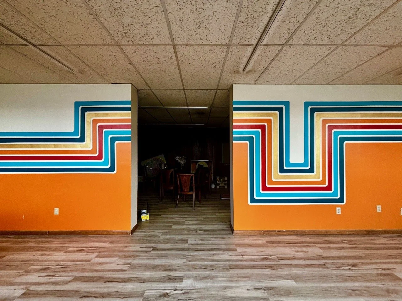 Colorful retro style wall mural with geometric lines in blue, red, yellow, and orange above a wooden floor, leading into a dark room with chairs.