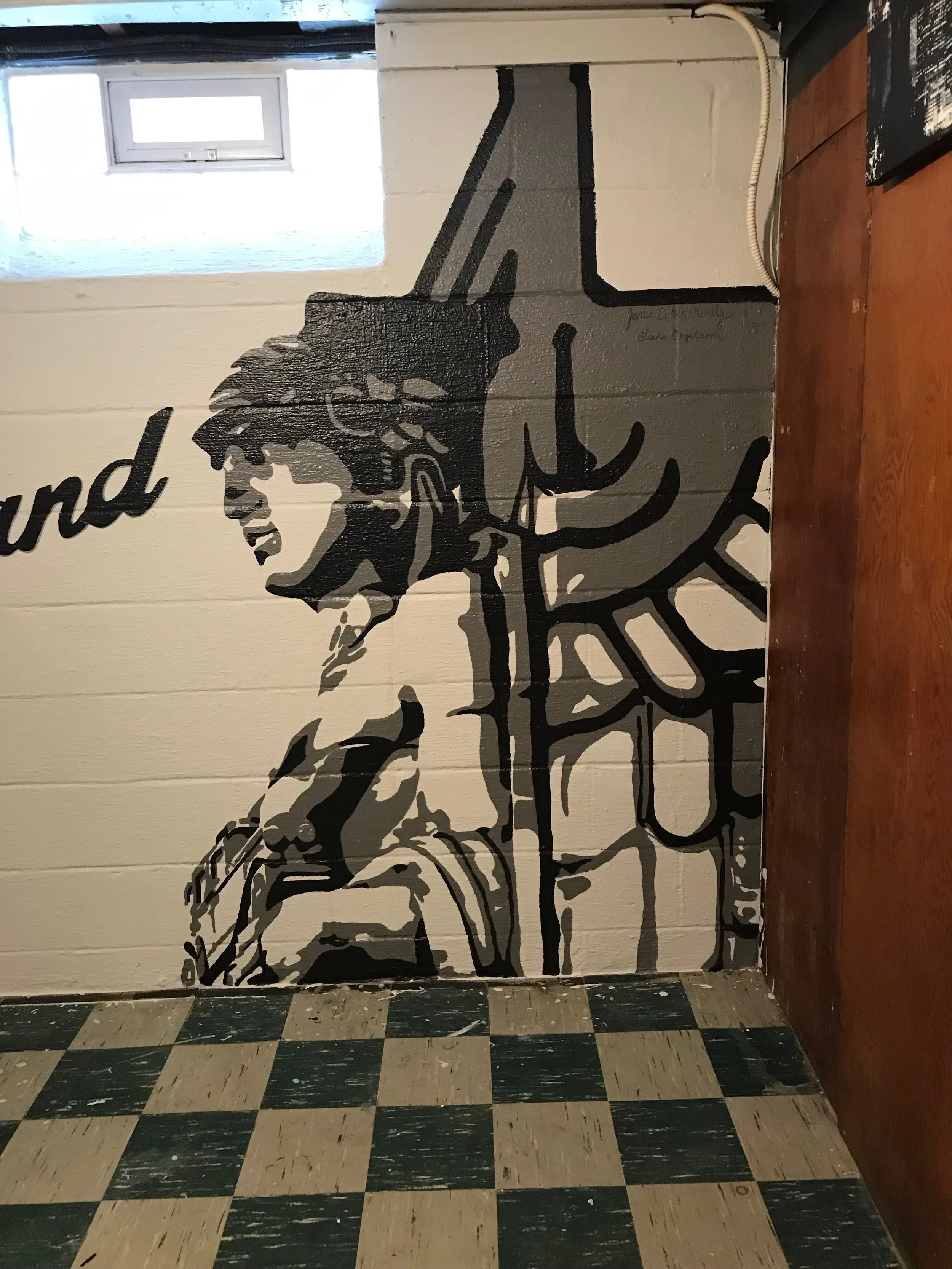 Wall mural of a black and white illustration of a woman wearing glasses, with text and artistic details. The mural covers a tiled wall near a small window, with a section of wooden counter and patterned tile floor visible.