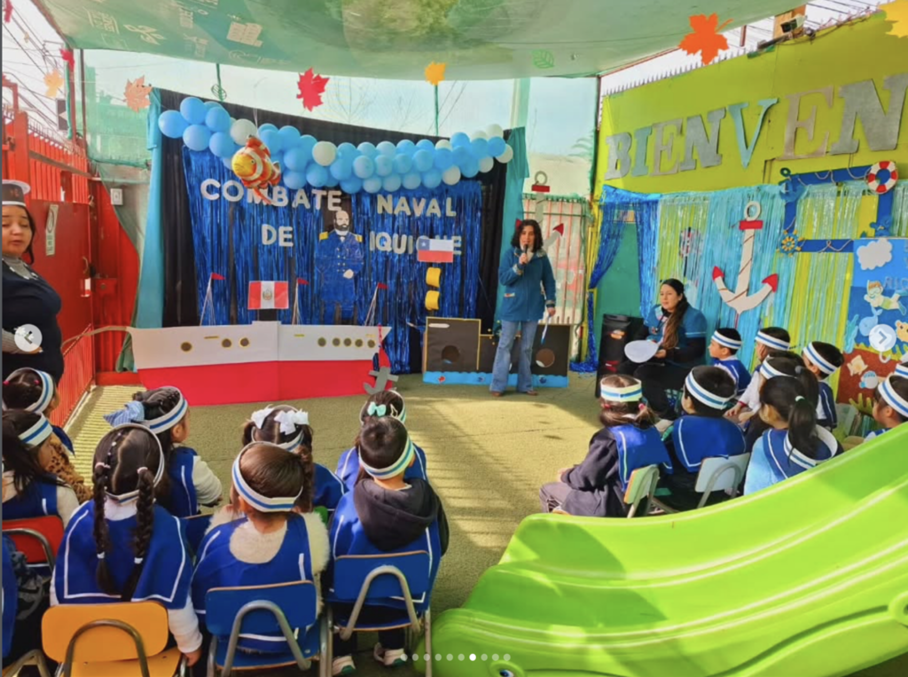 Niños en uniforme azul asistiendo a un evento escolar con decoración náutica, incluyendo globos azules y blancos, y un cartel que dice 'COMBATE NAVAL DE AQUÍ'. Hay un área de juegos de tobogán verde en primer plano y personas con vestimenta en unifor