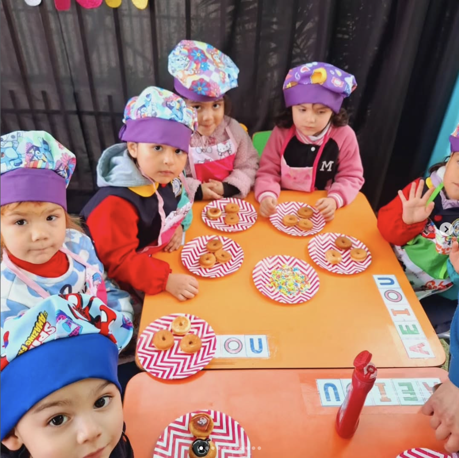 Niños en una celebración infantil con gorros de colores y personajes de dibujos animados, sentados en una mesa con platos de donas decoradas y letras del abecedario en el borde de la mesa.