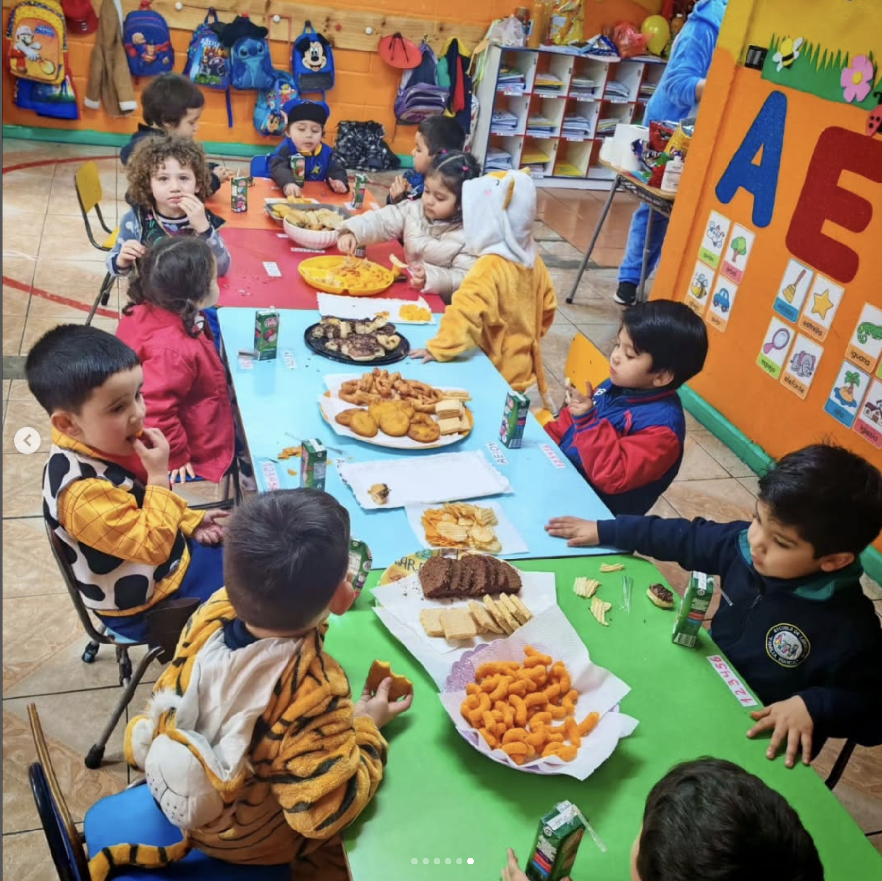 Niños en una celebración en la escuela, sentados en mesas con comida y bebidas, en un salón decorado con colores vivos y dibujos.