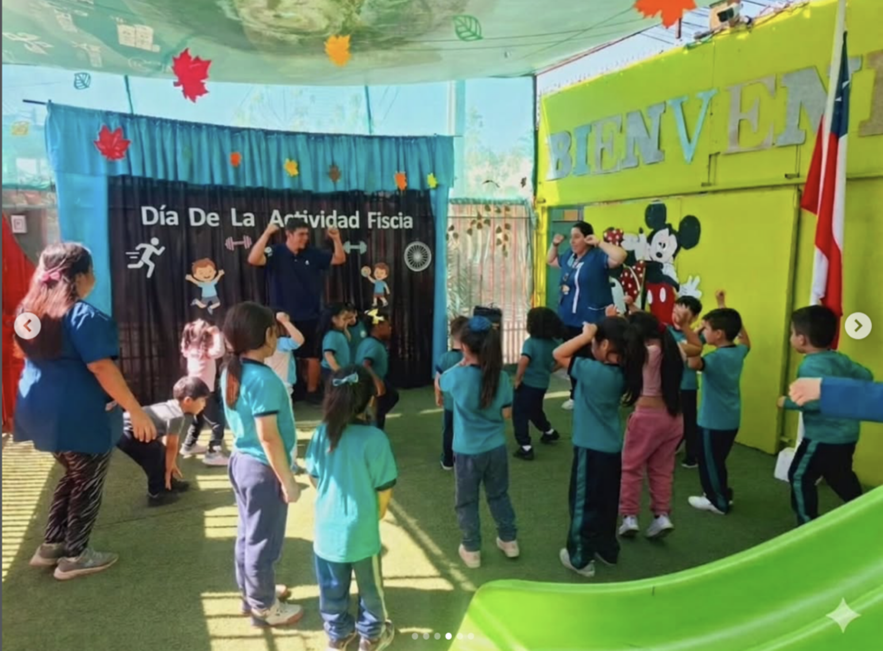 Niños participando en una actividad de ejercicio en un salón decorado con un cartel que dice 'Día de la Actividad Física' y una pared con el saludo 'Bienvenido', mientras un instructor los guía en una rutina de ejercicio.