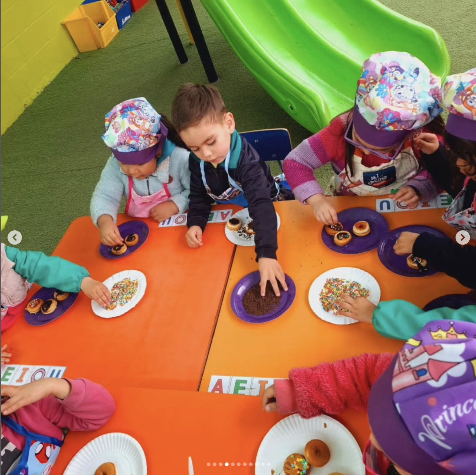 Niños pequeños en una mesa jugando con donuts decorados con chocolates y confites, en un área de juegos infantil con césped y resbaladilla verde.