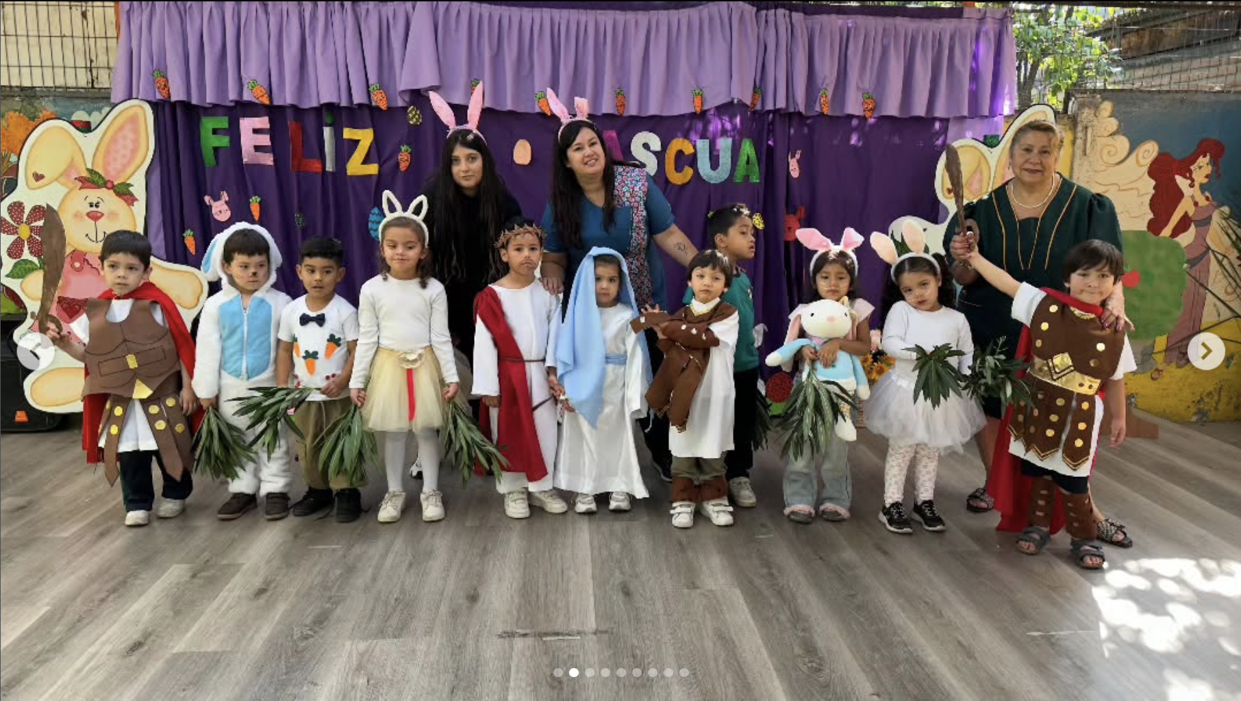 Grupo de niños disfrazados de personajes de la historia de Pascua, junto a dos adultas en un evento escolar, en un fondo decorativo con letras que dicen 'Feliz Pascua' y dibujos de conejos y otros símbolos de Pascua.