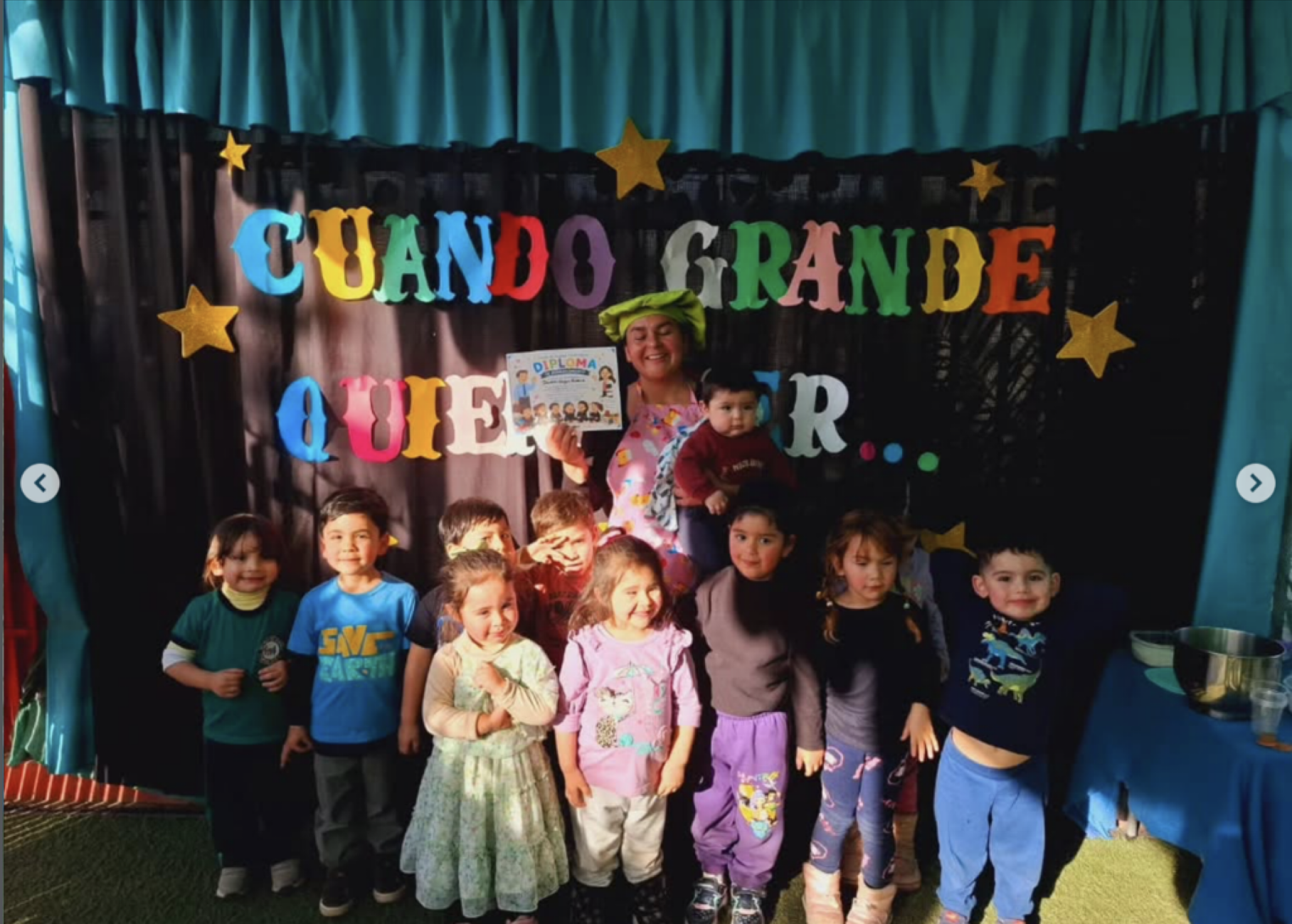 Grupo de niños y una mujer con gorro verde en celebración escolar con cartel que dice "Cuando grande quiero ser" y decoraciones de estrellas en el fondo.