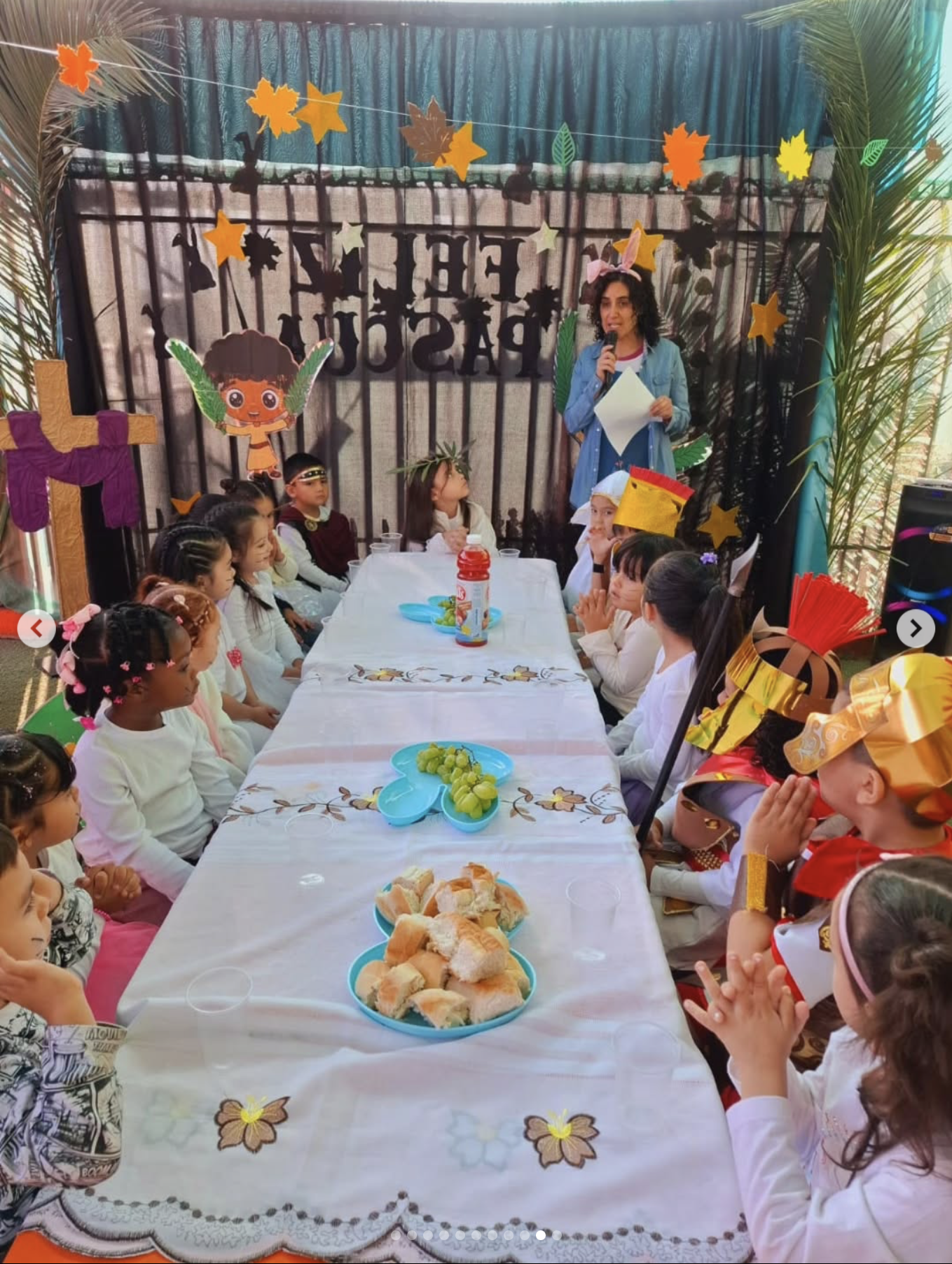 Niños vestidos con disfraces religiosos y tradicionales en una celebración, sentados alrededor de una mesa con comida, con decoraciones como hojas, estrellas y personajes de dibujos en el fondo.