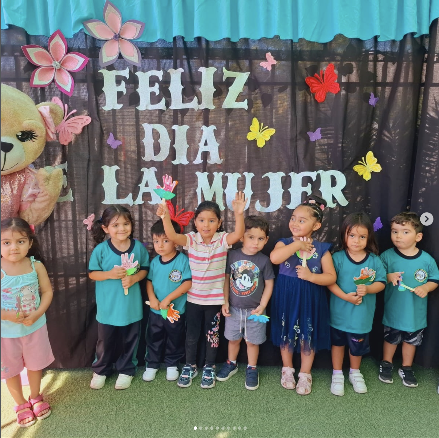 Niños y niñas con camisetas azules, algunos con accesorios y algunos con manos levantadas, en un evento escolar por el Día de la Mujer, con decoración de mariposas, flores de colores y un cartel que dice 'Feliz Día de la Mujer'.