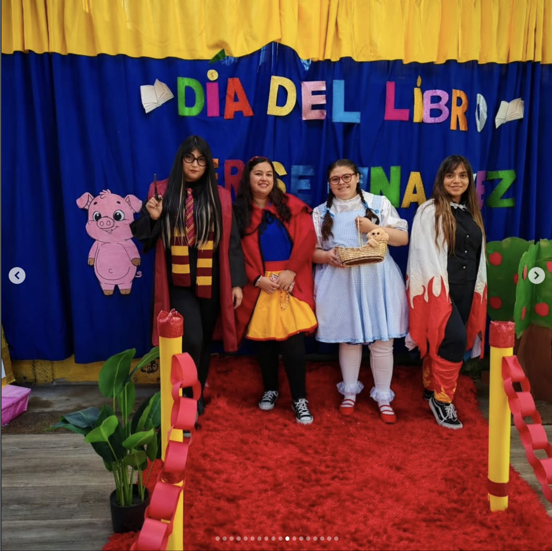 Grupo de personas disfrazadas en una celebración del día del libro, con decoraciones coloridas y personajes de dibujos animados, en un escenario con cortinas azules y fondo amarillo.