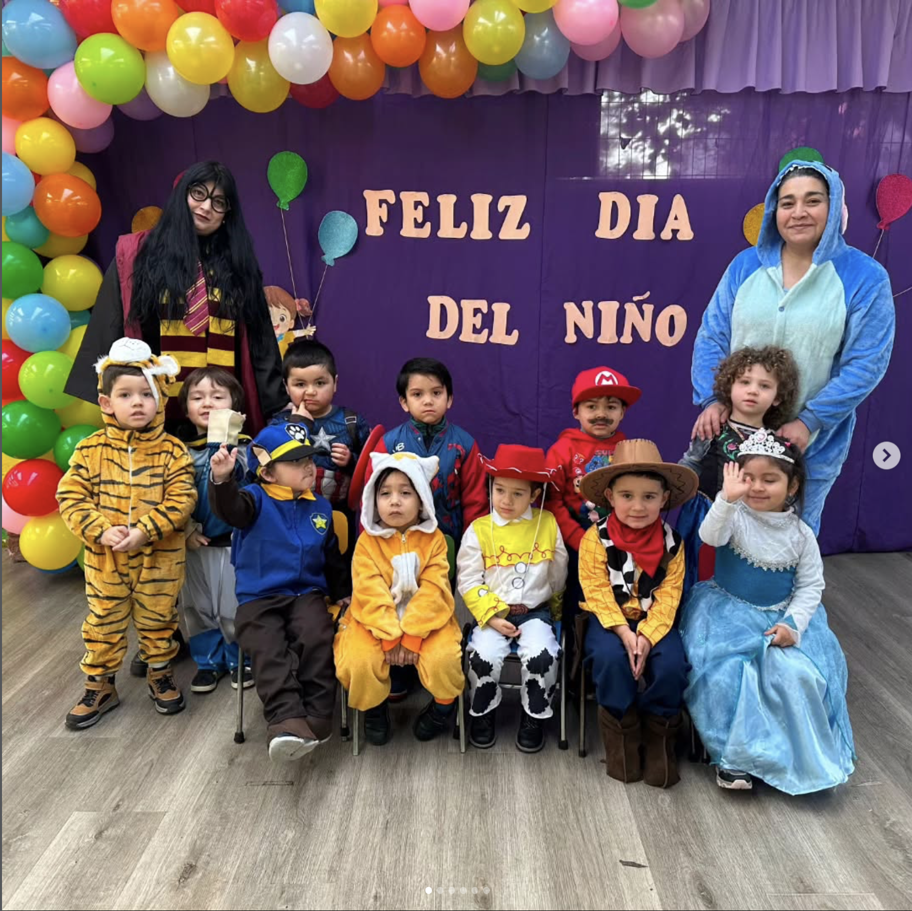 Niños disfrazados de animales y personajes, acompañados por dos mujeres, en una celebración por el día del niño, con globos de colores y un cartel que dice 'Feliz Día del Niño' en un fondo púrpura.