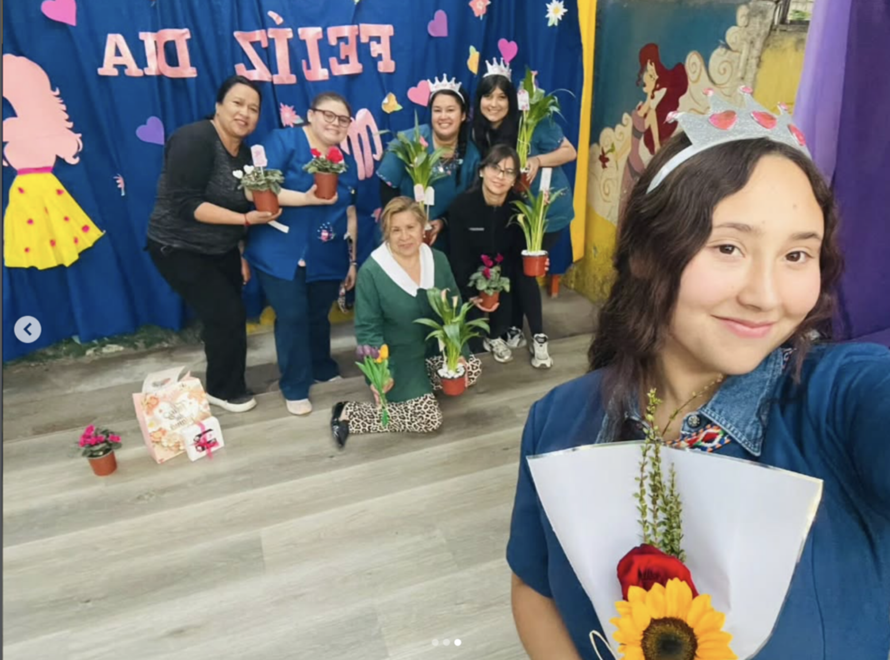 Grupo de mujeres en una celebración del Día de la Madre. Algunas llevan coronas y sostienen plantas en macetas. La decoración incluye un fondo azul con corazones y figuras femeninas, y en el suelo hay flores y regalos.