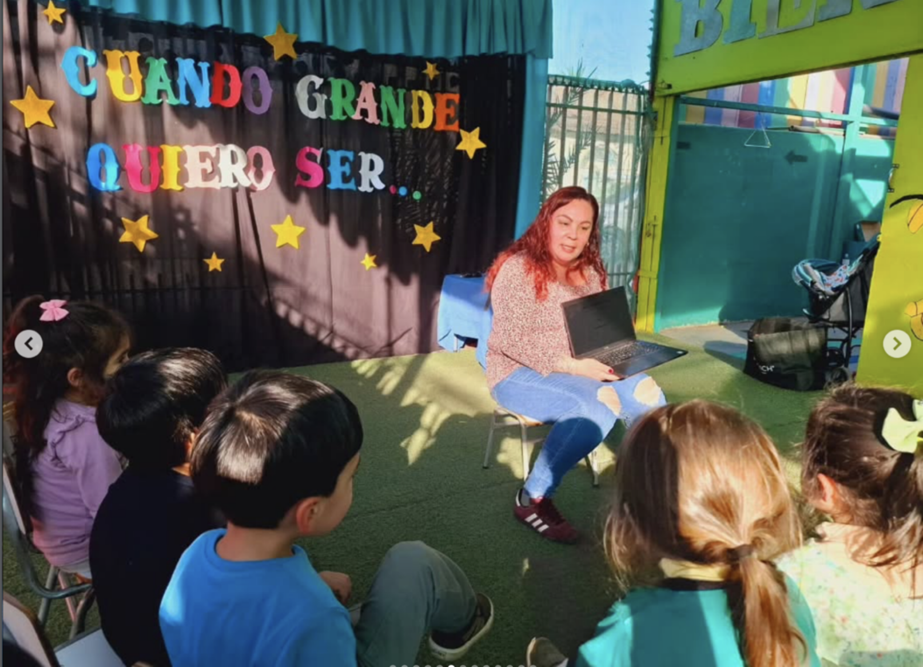 Una mujer con cabello rizado rojo dando una charla a un grupo de niños en un espacio decorado con letras y estrellas de colores que dice "Cuando grande quiero ser" en un entorno de estilo tropical con vista a la ciudad al fondo.