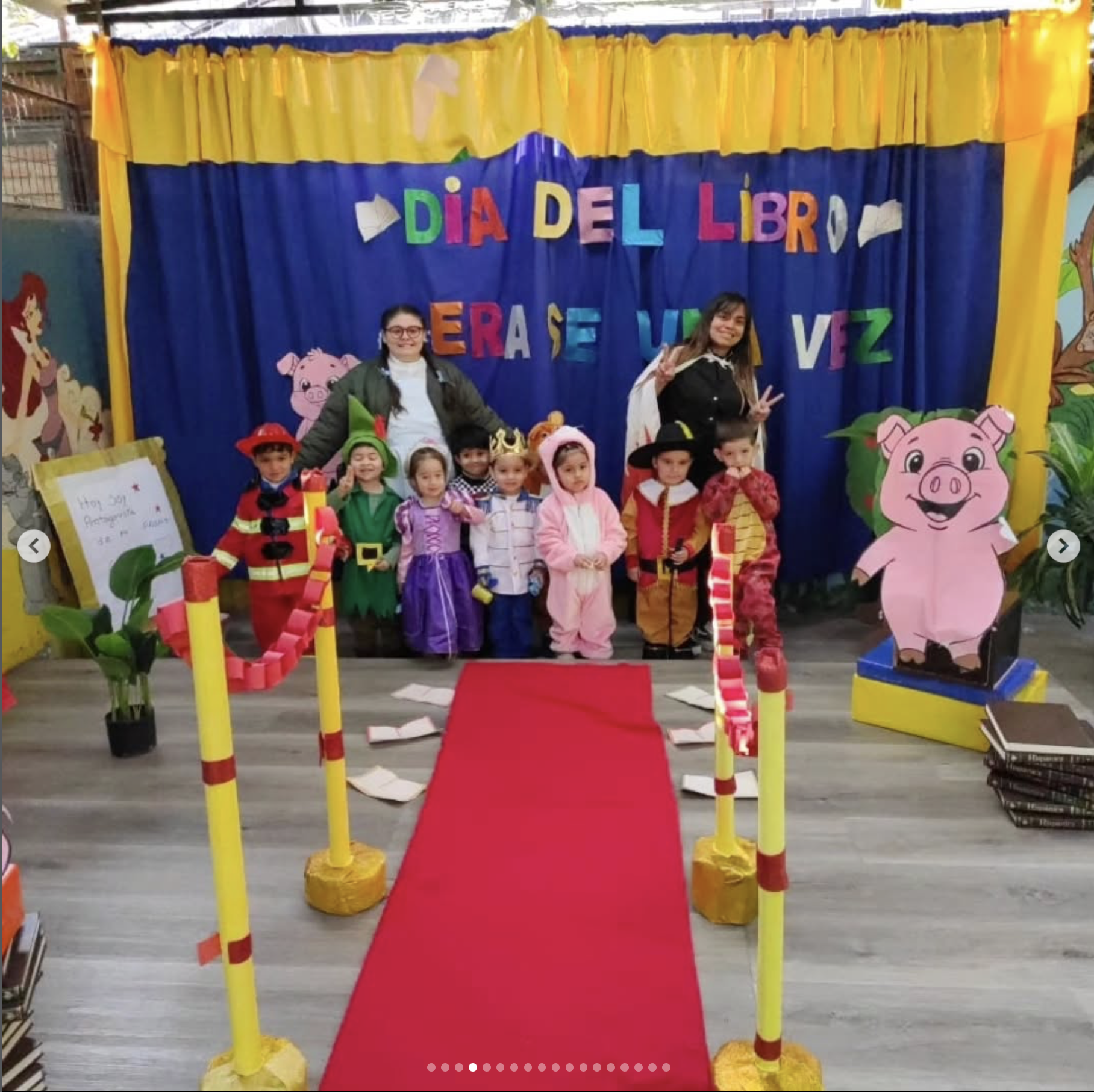 Escenario decorado para celebrar el Día del Libro, con banderines coloridos, una alfombra roja y figuras de personajes infantiles, con niños y adultos disfrazados en el centro.