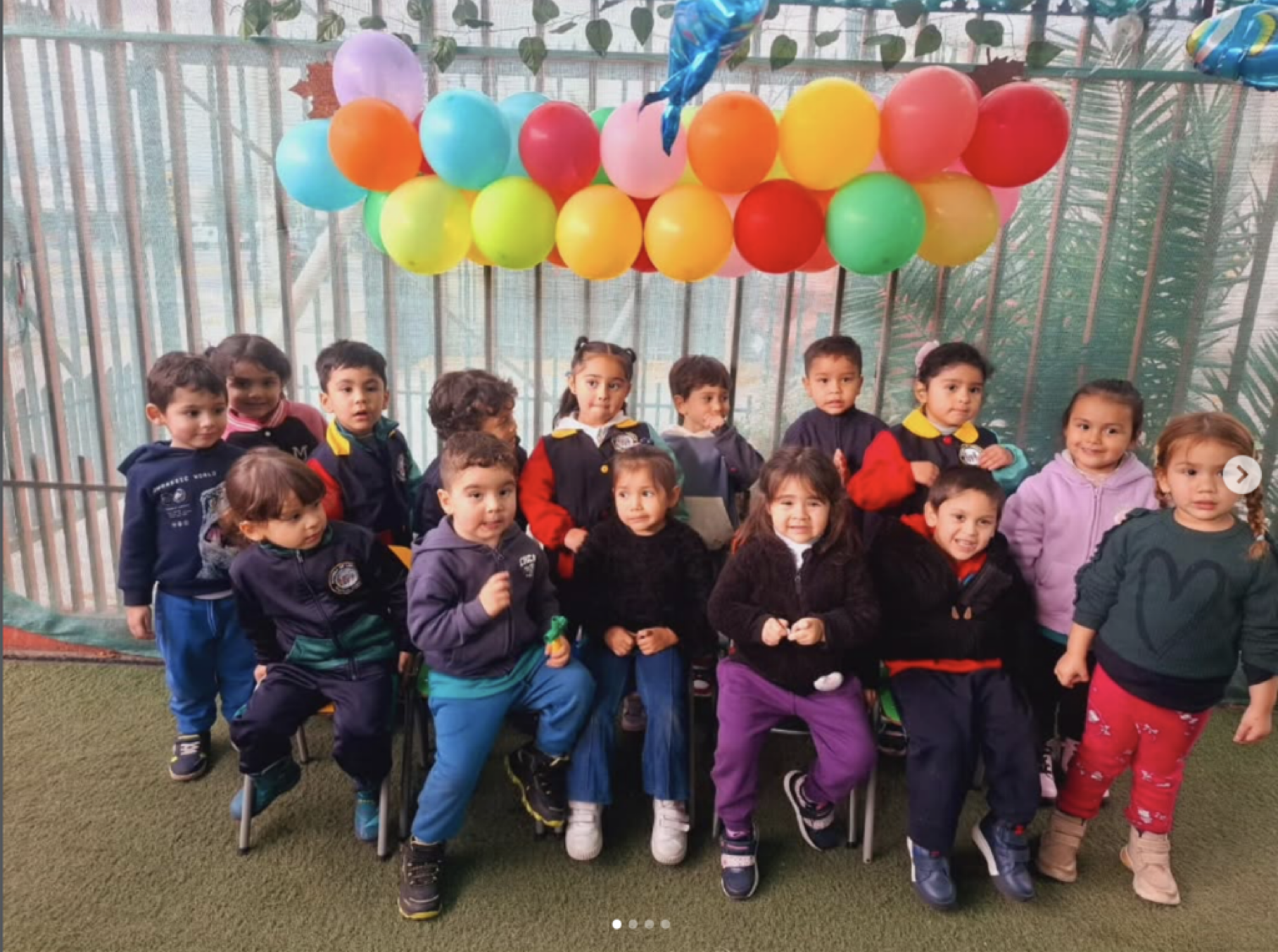 Grupo de niños y niñas pequeños en una celebración, con globos de colores en el fondo, en un espacio cubierto con malla de protección.