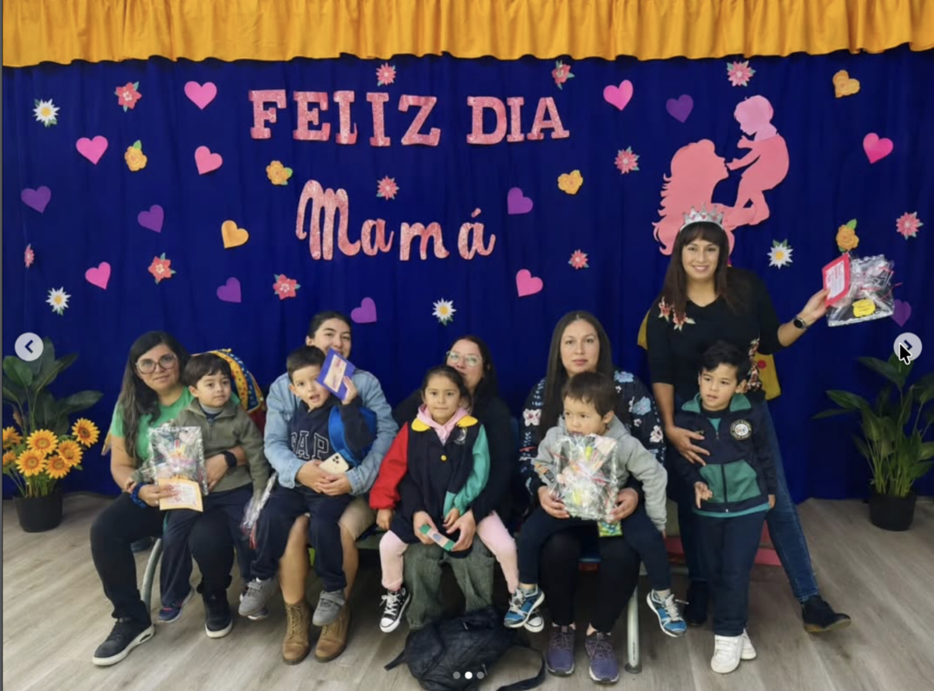Grupo de personas, niños y adultos, posando frente a un fondo decorado con corazones, flores y la frase 'Feliz Día Mamá'.