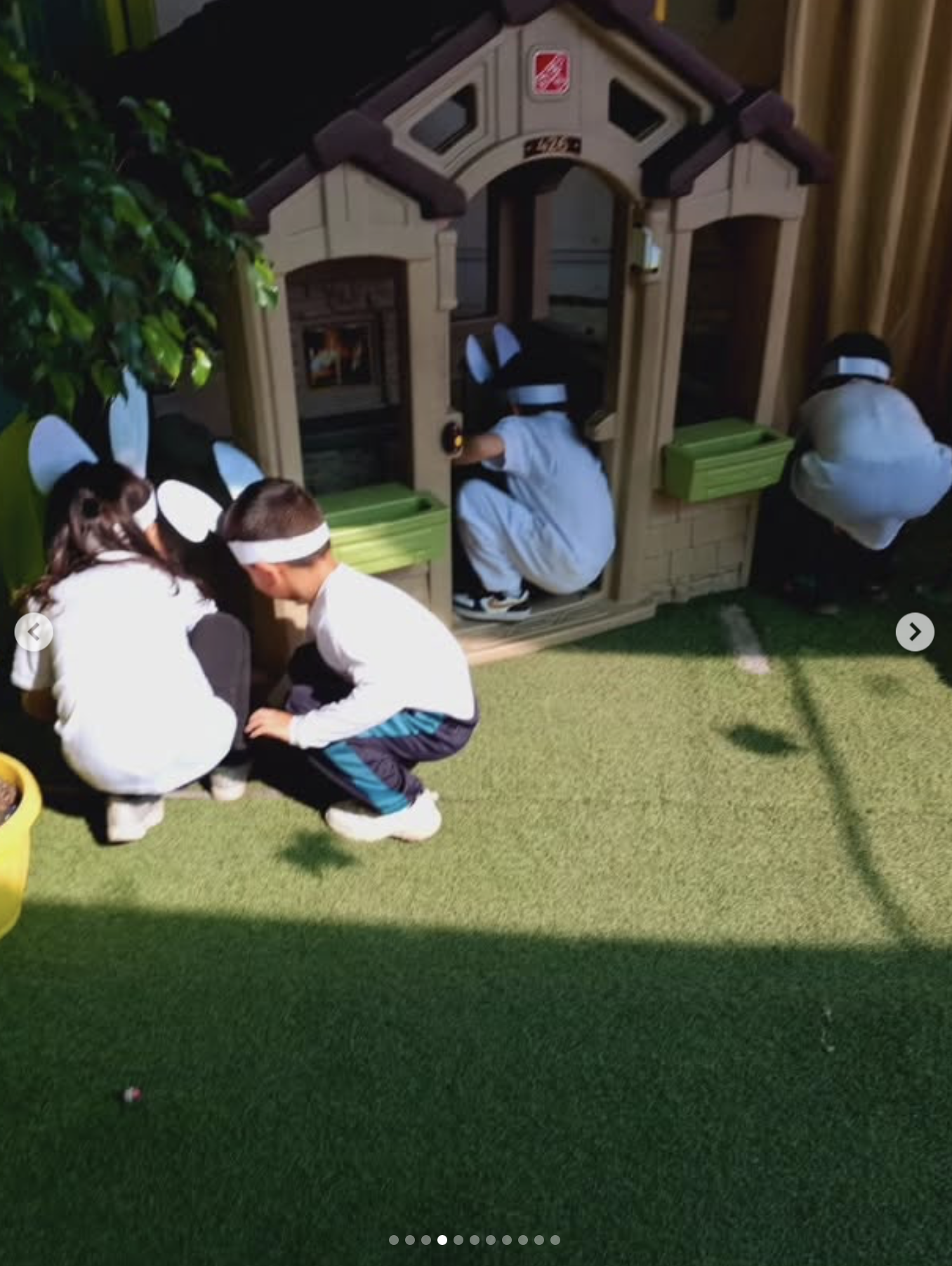 Niños disfrazados de conejos jugando en un jardín con césped artificial, cerca de una casita de madera con chimenea y plantas decorativas.