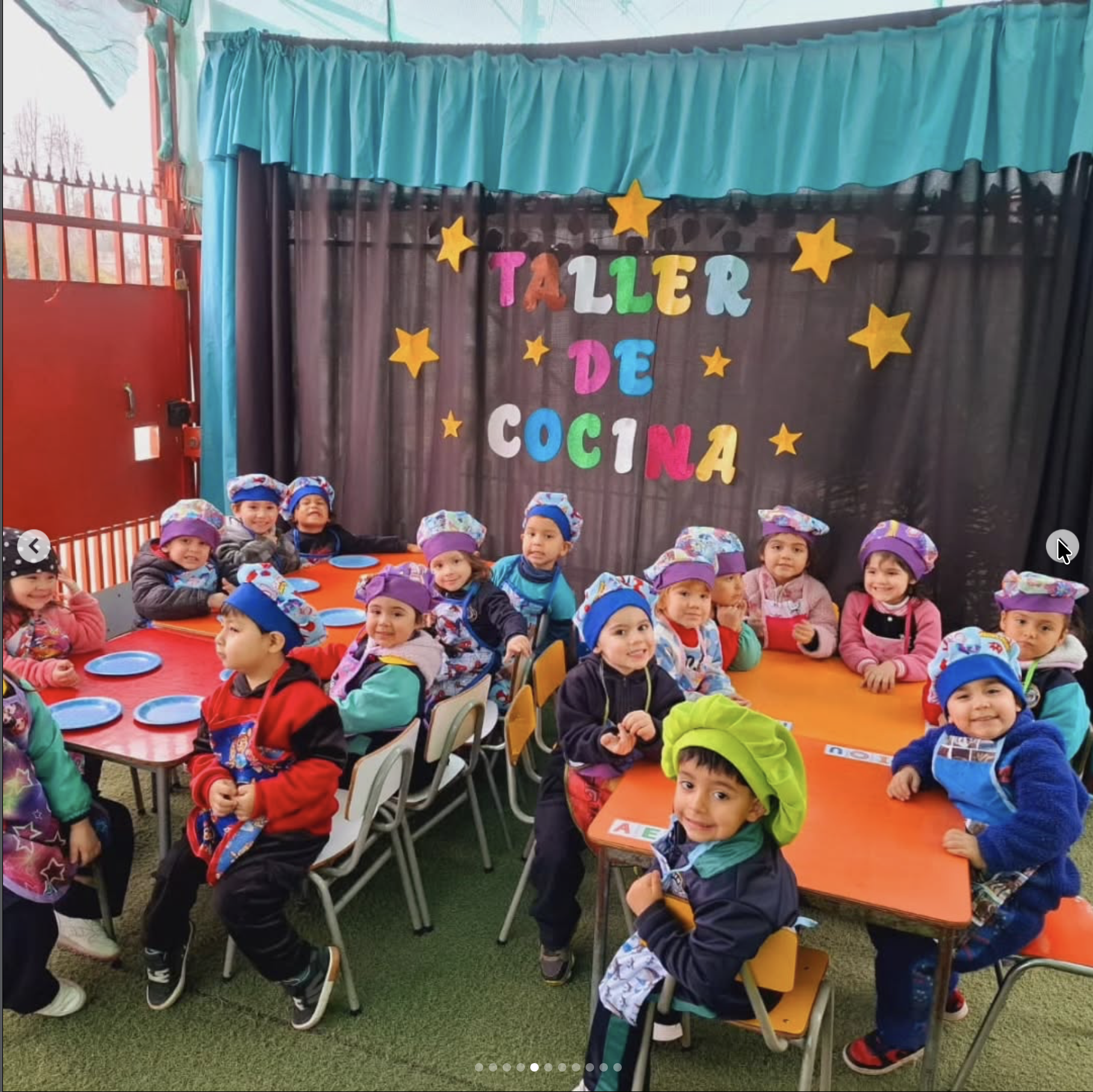 Niños en un taller de cocina, usando delantales y gorros, sentados en mesas con platos y cubiertos, en un ambiente decorado con estrellas y letras de colores que dice 'Taller de Cocina'.