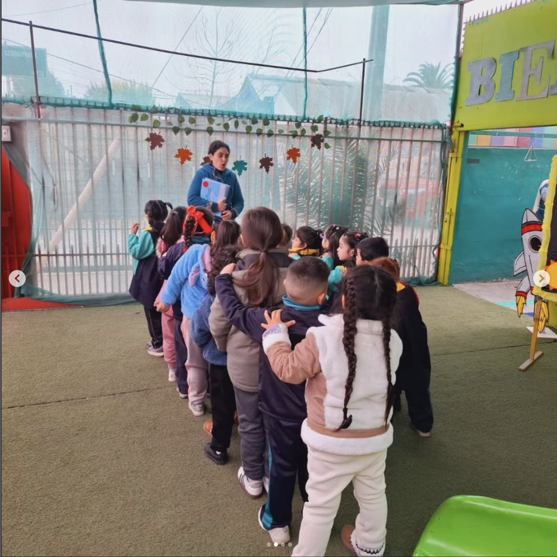 Grupo de niños en fila en un espacio cubierto, siendo supervisados por una mujer con documentos en mano, en un ambiente escolar infantil decorado con hojas y temática de verano o escuela.