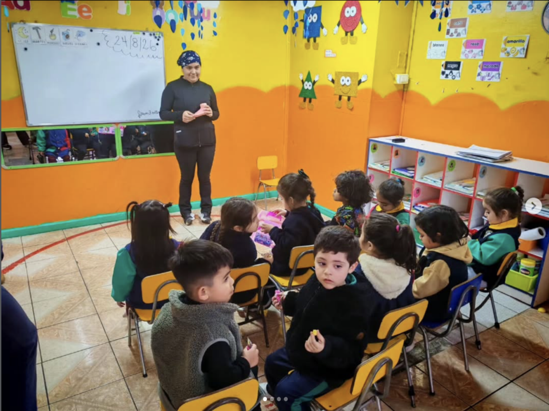 Niños en un aula escolar con una maestra de pie frente a la pizarra. La sala está decorada con personajes coloridos en las paredes y estantería con libros y materiales educativos.