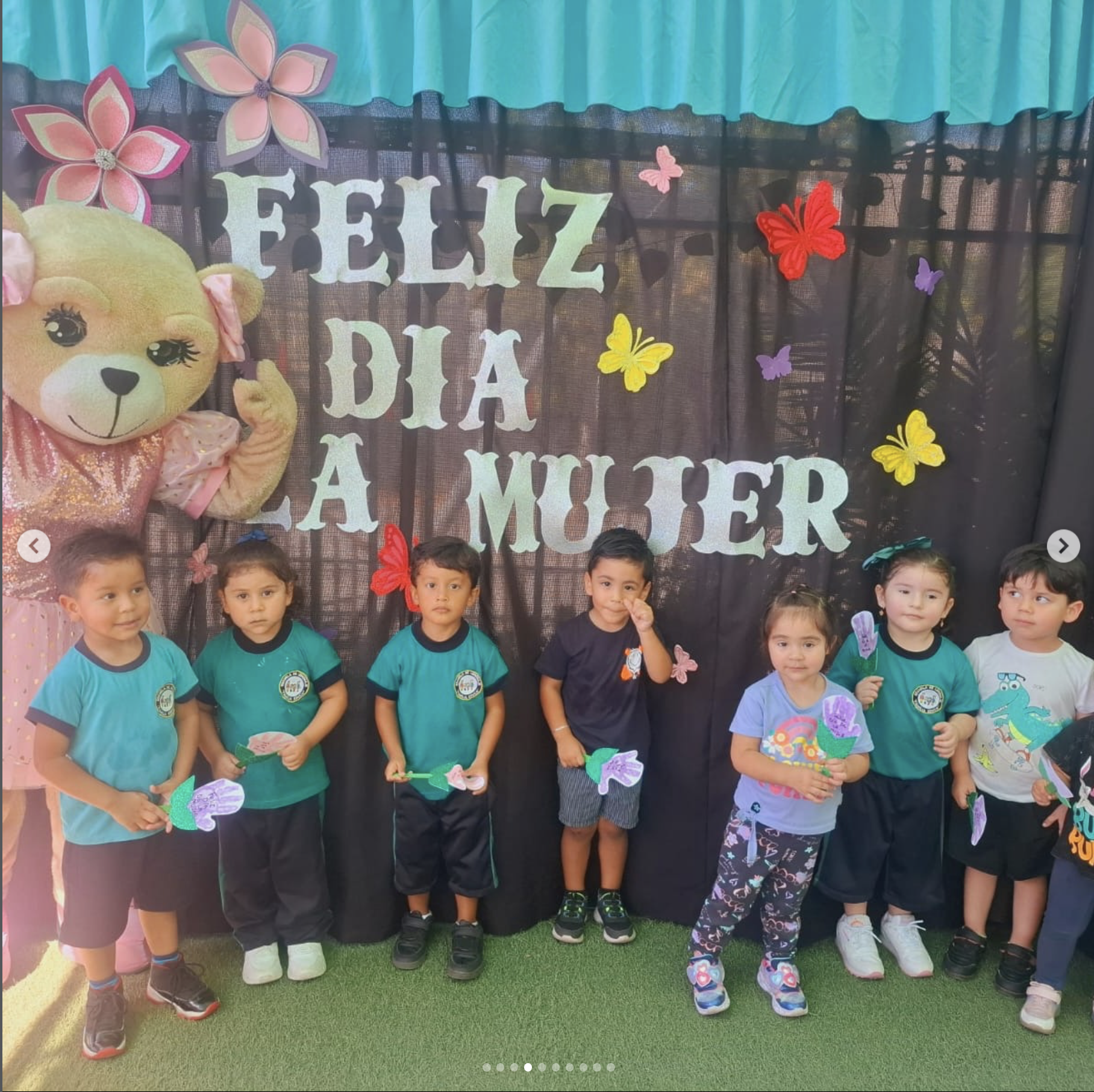 Grupo de niños posando frente a un fondo decorado con letras que dicen 'Feliz Día La Muler'. Un oso de felpa grande con vestido rosa y la cara visible en la esquina izquierda está acompañado por las niñas y niños. Las niñas sostienen mariposas de pap