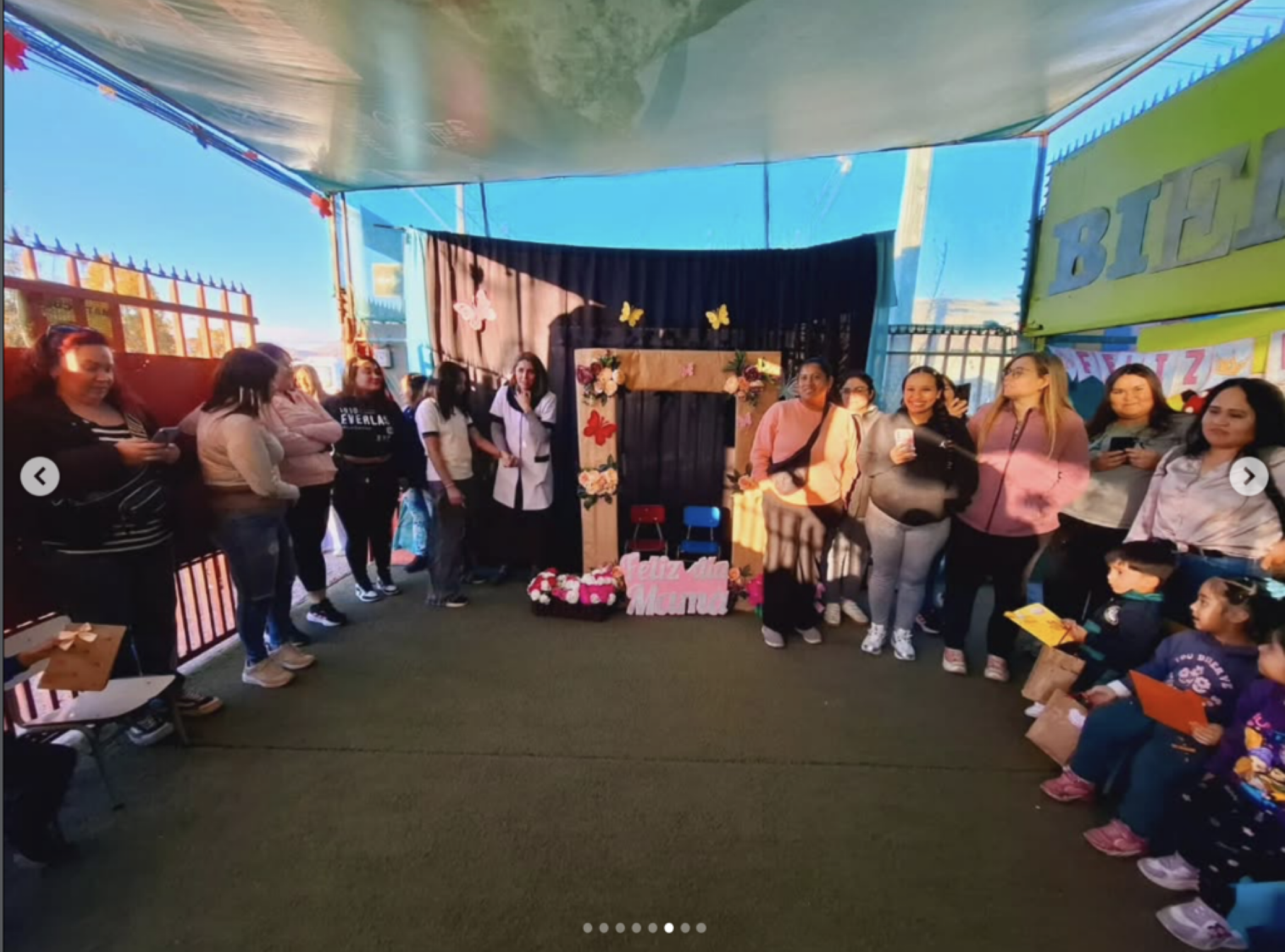 Grupo de personas, en su mayoría mujeres, de pie en un espacio cerrado decorado con mariposas y flores, participando en una celebración por el Día de la Madre, con un cartel que dice "Feliz Día Madre".