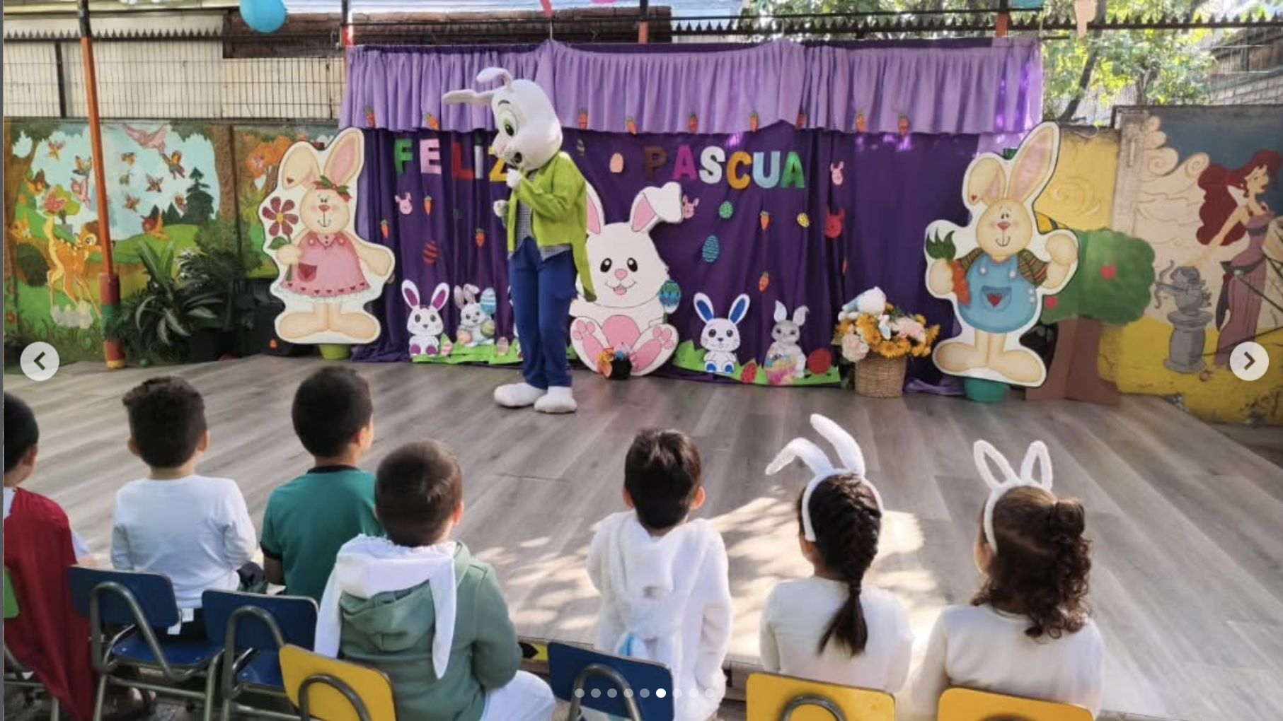 Niños asistiendo a una obra de teatro con personajes de conejo en un escenario decorado con dibujos de Pascua y flores.
