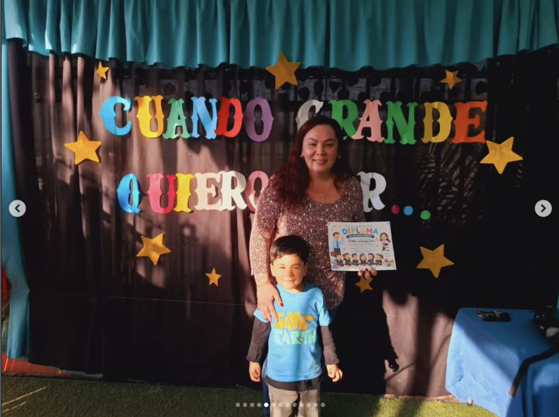 Niña y niño posando en una celebración escolar con cartel que dice 'Cuando grabe, quiero', y una mujer sosteniendo un diploma, decoraciones con estrellas doradas.