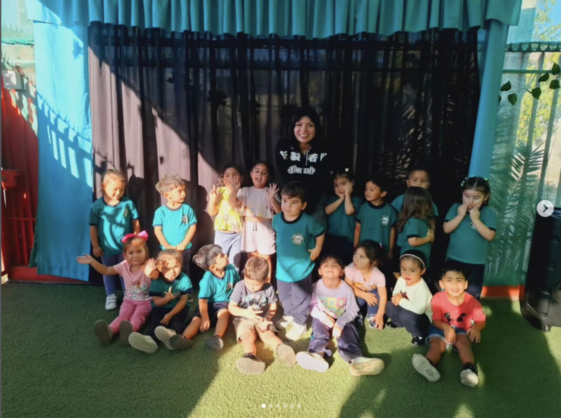 Grupo de niños y una adulta en un espacio cubierto con cortinas de colores y cortinas negras, en un ambiente soleado con plantas verdes en el fondo, todos posando para la foto.
